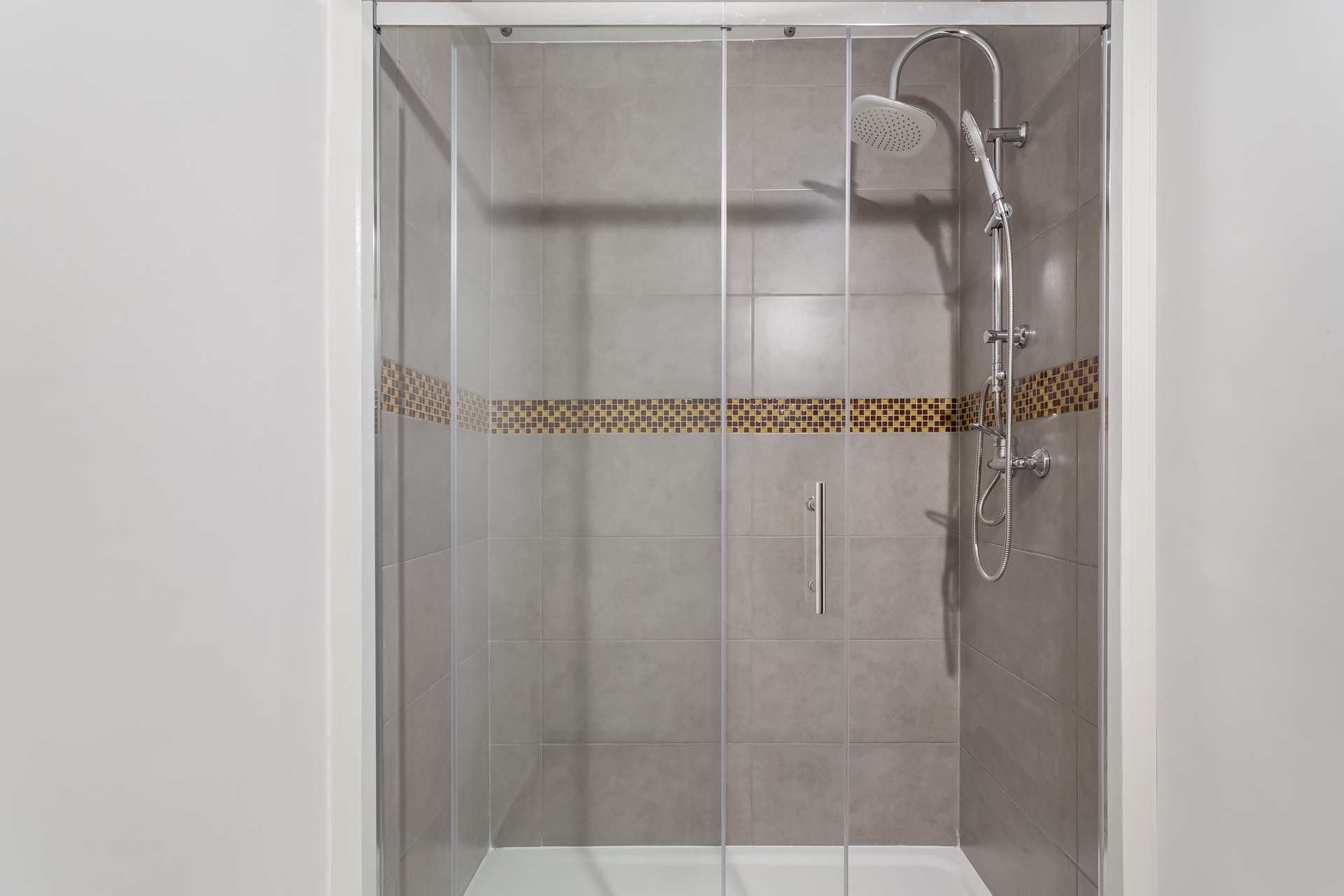 Dusche mit Glas-Schiebetür, grauen Fliesen und einer Zierleiste aus Mosaikfliesen.