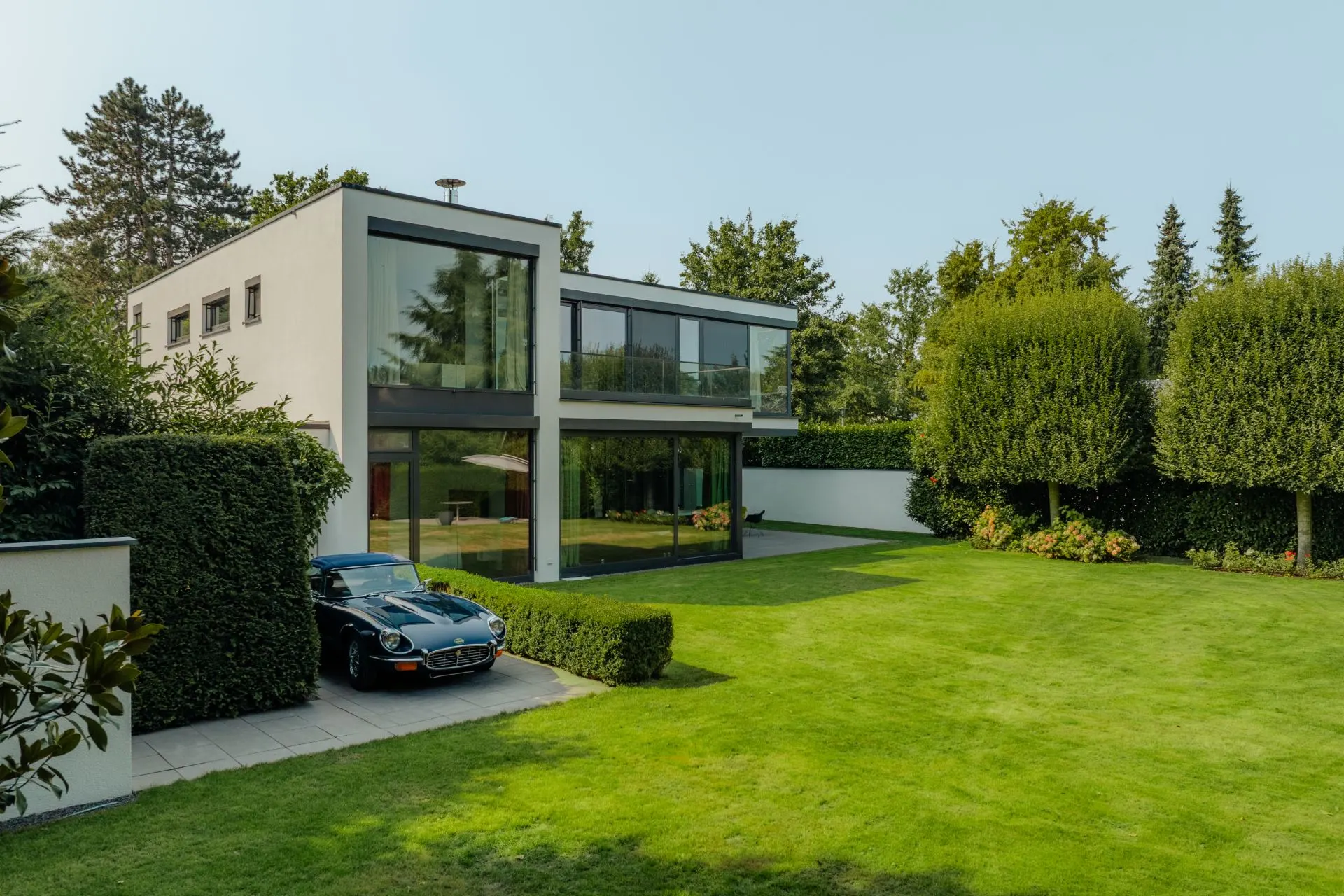 Modernes zweistöckiges Haus mit großen Fenstern und gepflegtem Garten, Auto steht vor dem Gebäude.