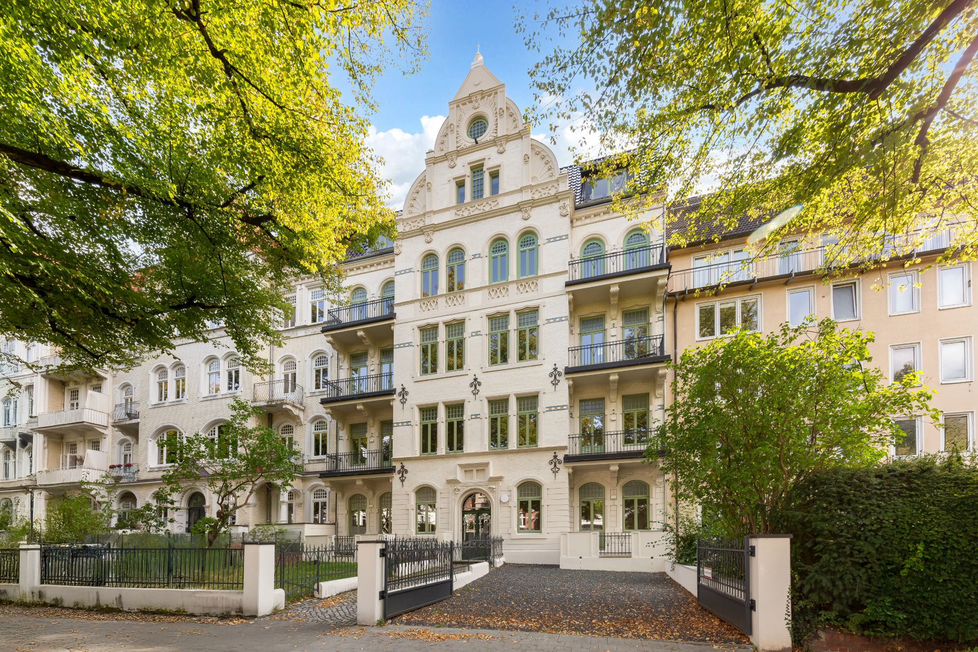Mehrstöckiges, helles Wohnhaus mit Balkonen, Garten und Einfahrt, umgeben von Bäumen.
