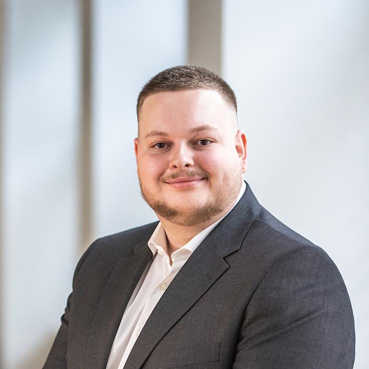 Ein Mann im Anzug steht vor einem hellen Hintergrund und lächelt in die Kamera.