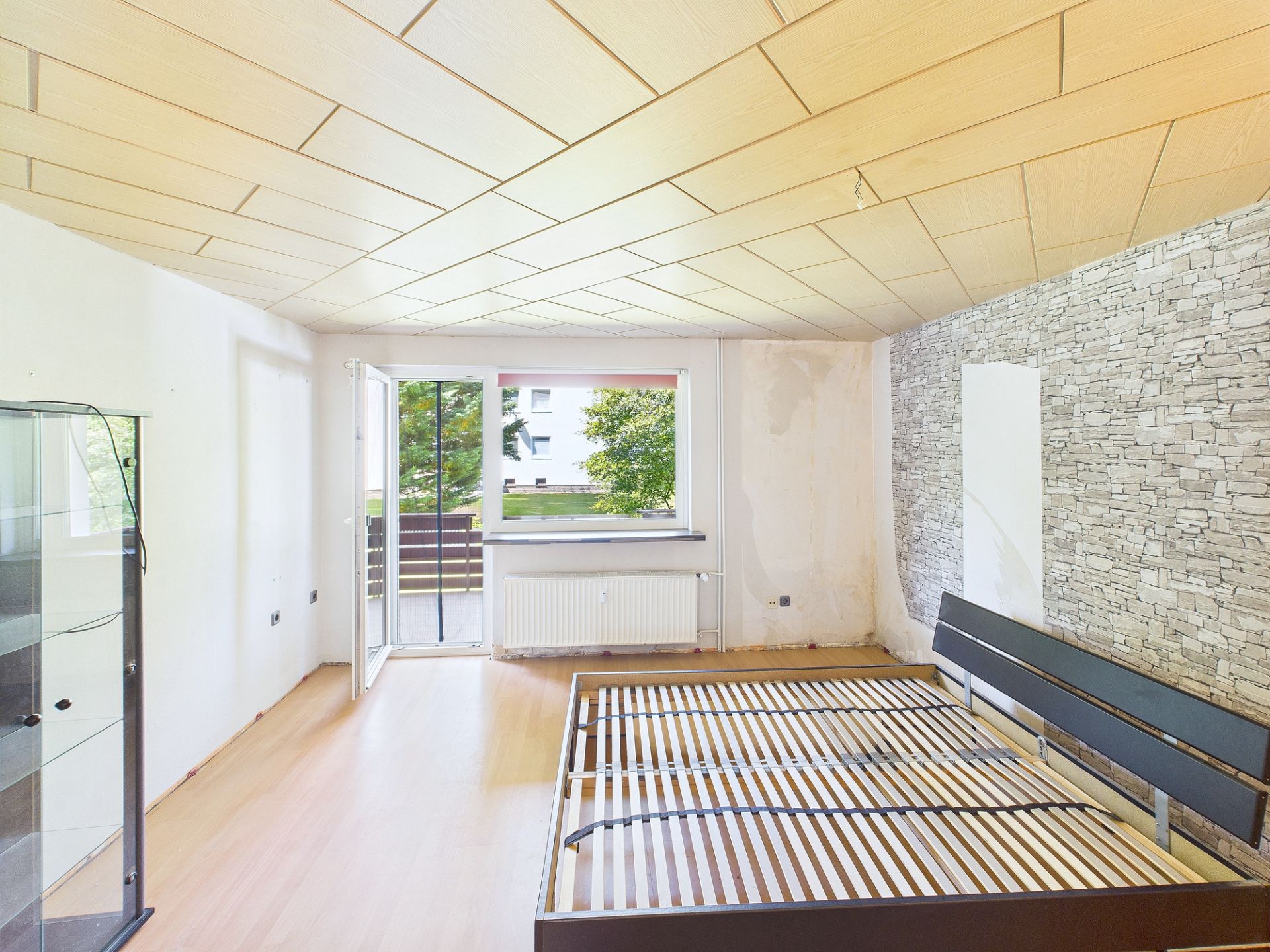 Helles Schlafzimmer mit leerem Bettgestell, Glasvitrine und Ausgang zum Balkon.
