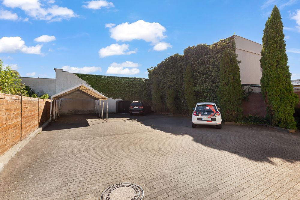 Gepflasterter Parkplatz mit zwei Autos, Holzzaun links und hohen Bäumen rechts.