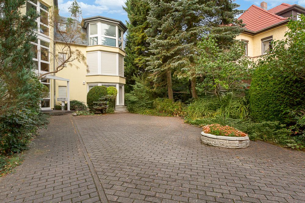 Gepflasterte Einfahrt mit Blumenbeet, Bäumen und gelbem Wohnhaus im Hintergrund.