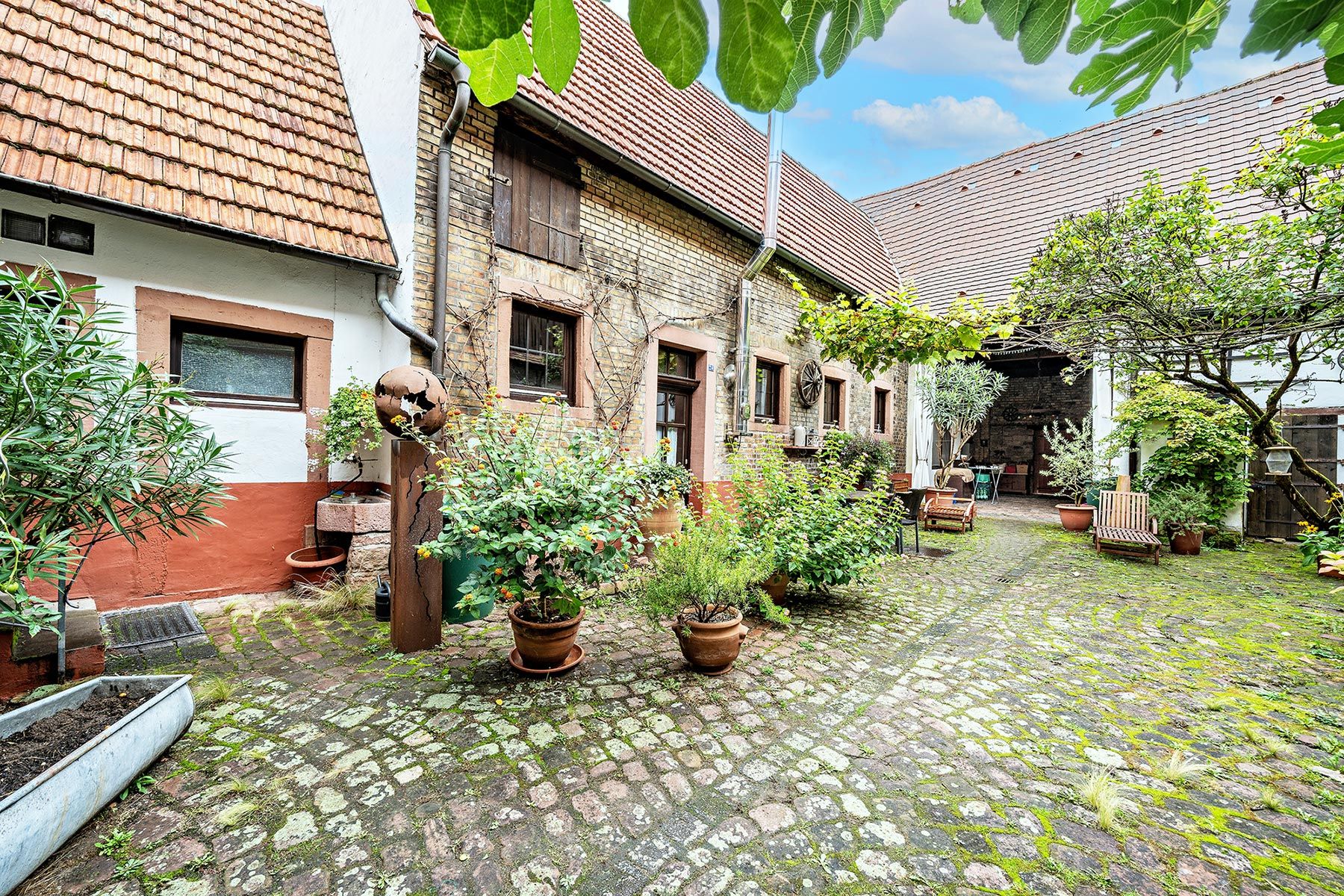 Gepflasterter Innenhof mit Pflanzen in Töpfen, Holzbänken und gemauerten Gebäuden.