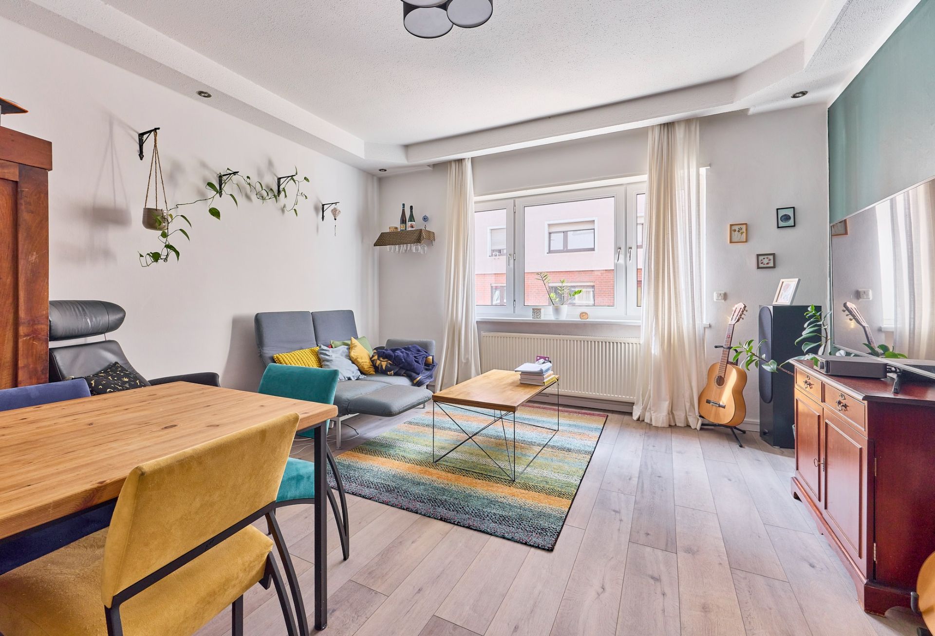 Helles Wohnzimmer mit Sofa, Holztisch, buntem Teppich und Gitarre neben dem Fenster.