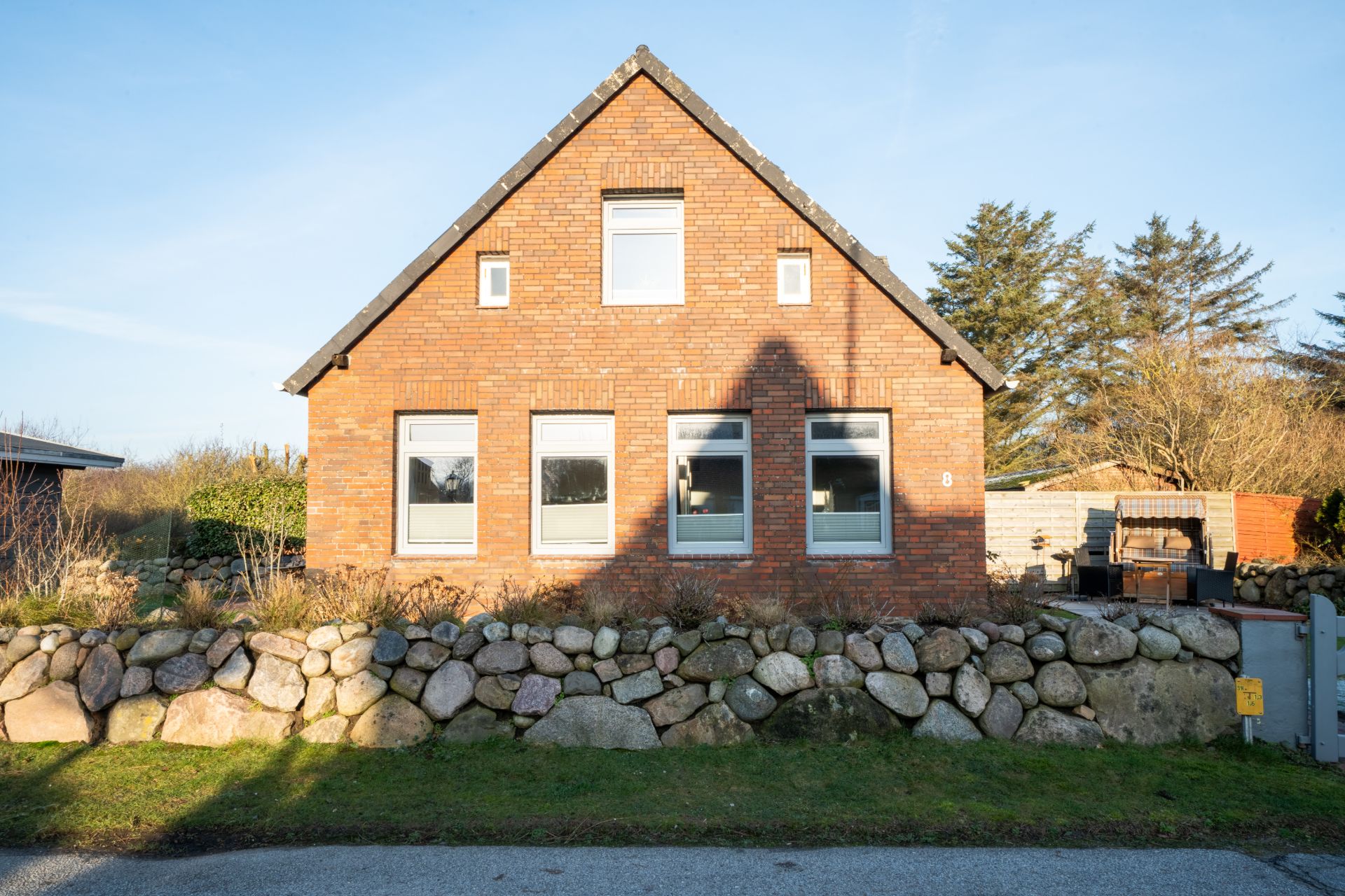 Backsteinwohnhaus mit Satteldach, drei großen Fenstern und Steinmauer im Vorgarten.