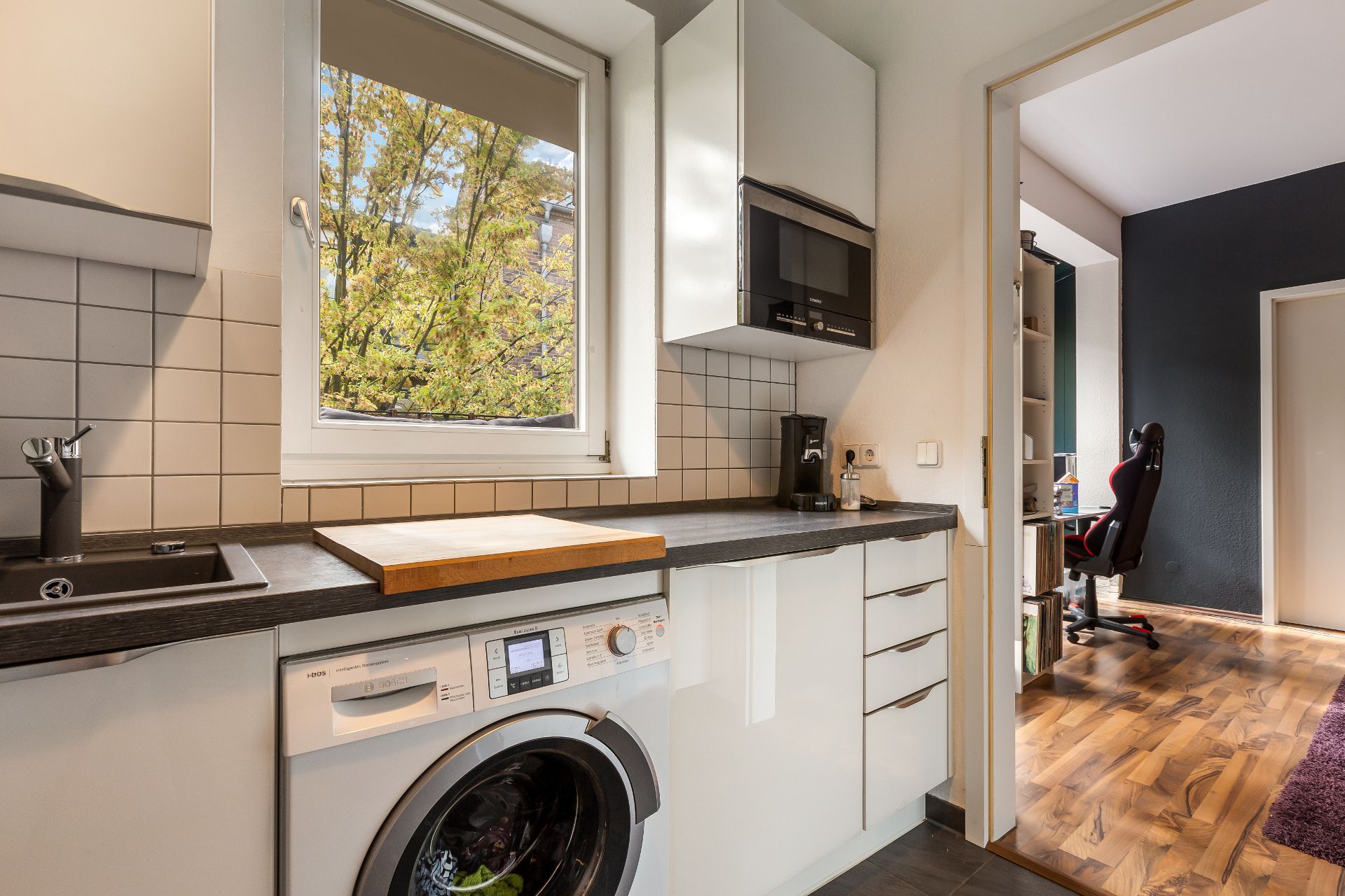 Moderne Küche mit Waschmaschine und Blick in ein Arbeitszimmer mit Schreibtischstuhl.