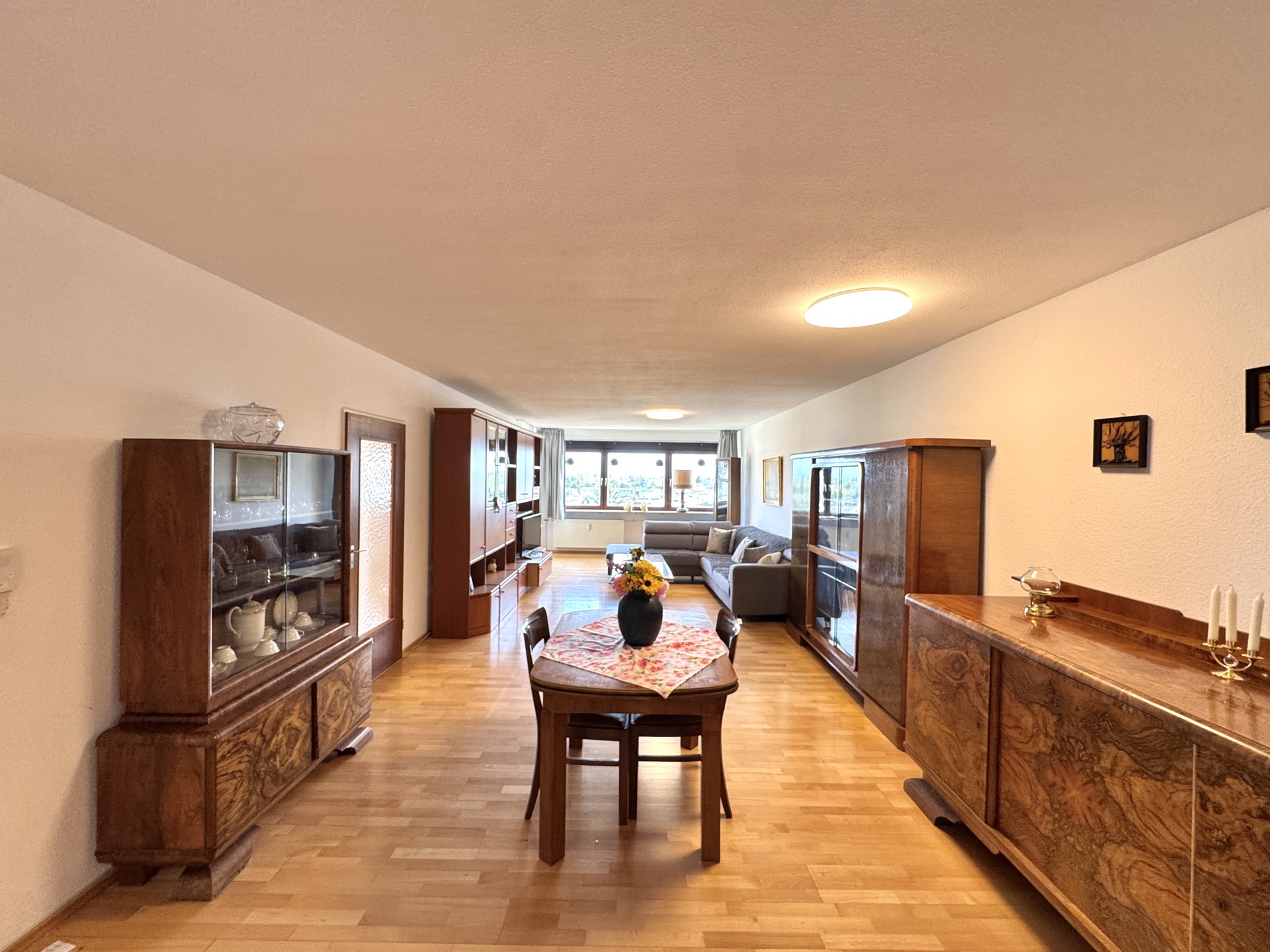 Helles Wohnzimmer mit Holzboden, Esstisch, Vitrinen, Sofa und großen Fenstern.