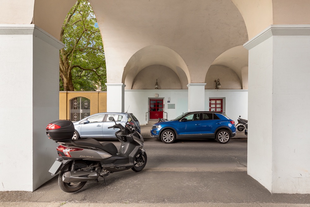 Motorrad und zwei Autos parken unter einem hellen Torbogen mit roten Türen im Hintergrund.