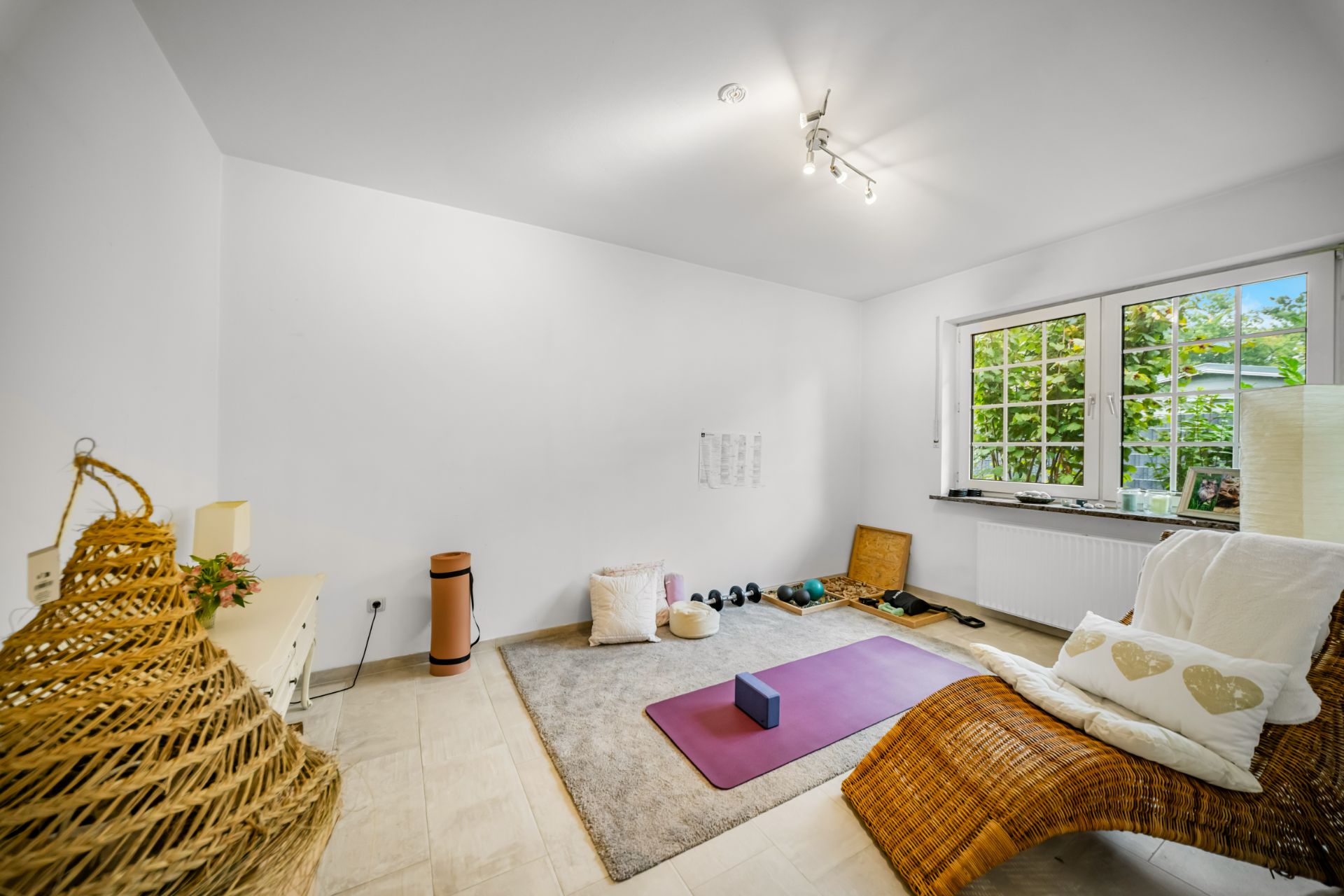 Heller Raum mit Yogamatte, Kissen, Hanteln und Rattanliege am Fenster.