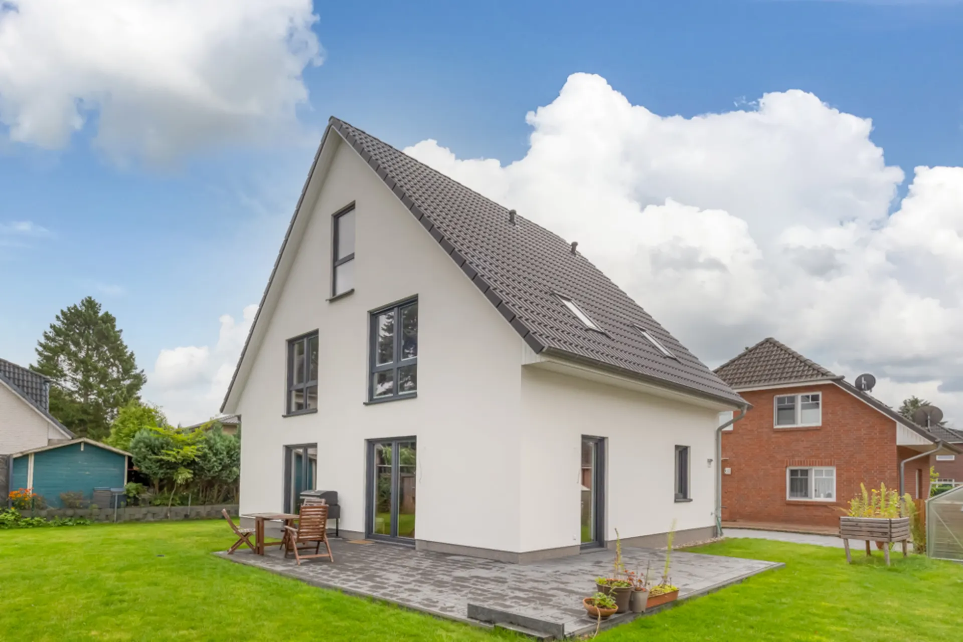Modernes Einfamilienhaus mit weißer Fassade, Terrasse und Garten, daneben weitere Häuser.