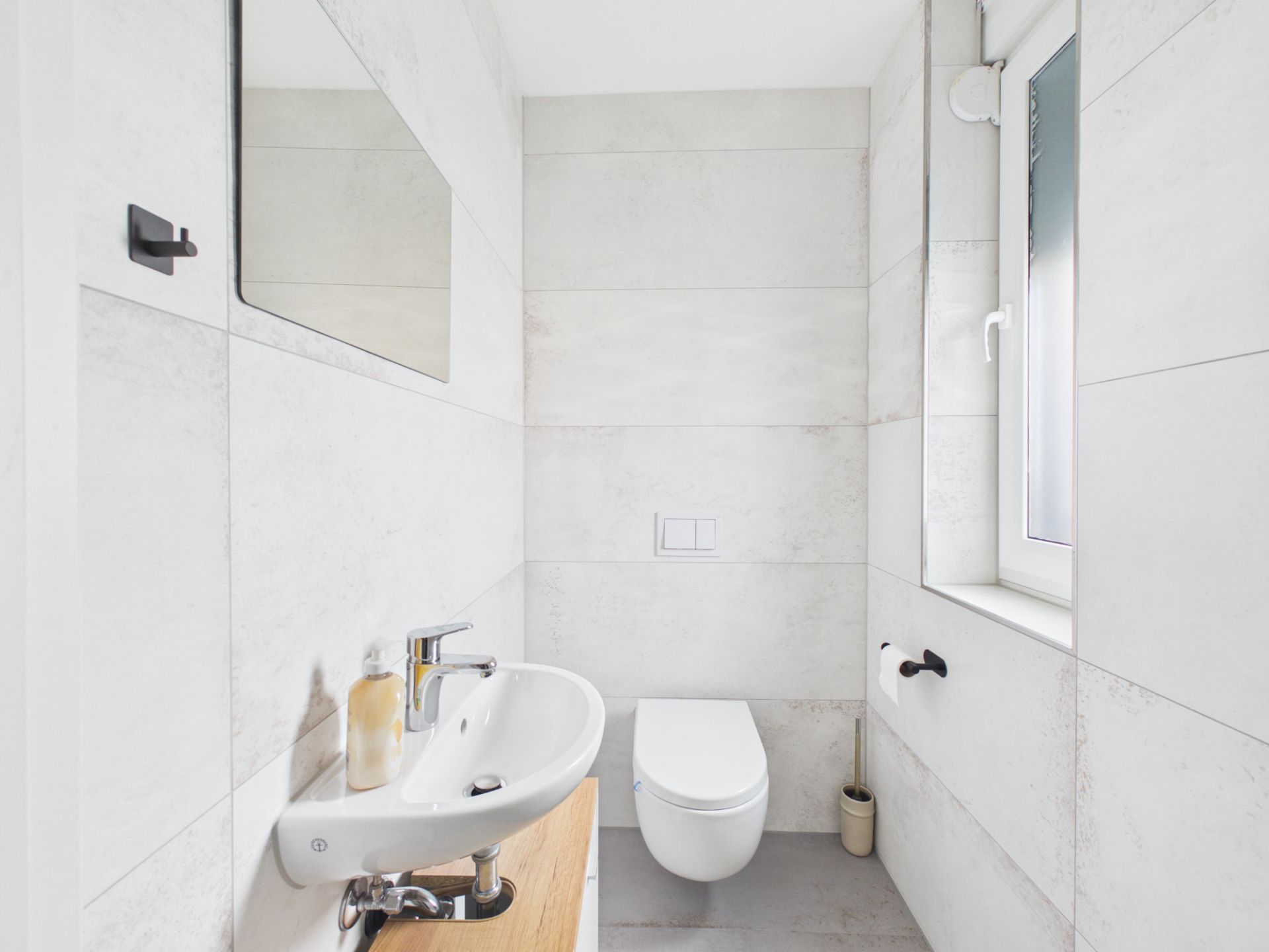 Helles Badezimmer mit Wandwaschbecken, Toilette, Spiegel und Fenster.