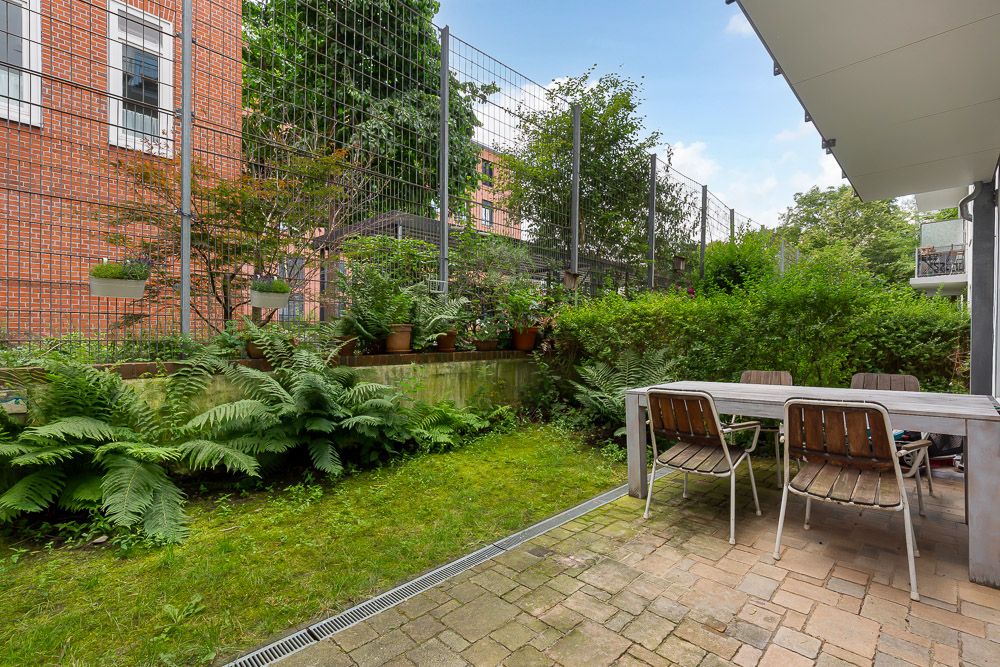 Kleiner Garten mit Terrasse, Tisch und vier Stühlen, umgeben von Pflanzen und Sträuchern.