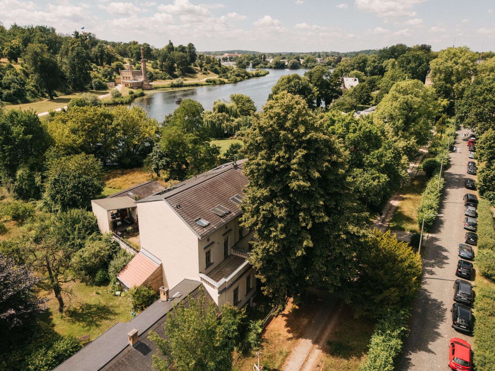 Wohnhaus mit Garten an einem Fluss, umgeben von vielen Bäumen und parkenden Autos auf der Straße.