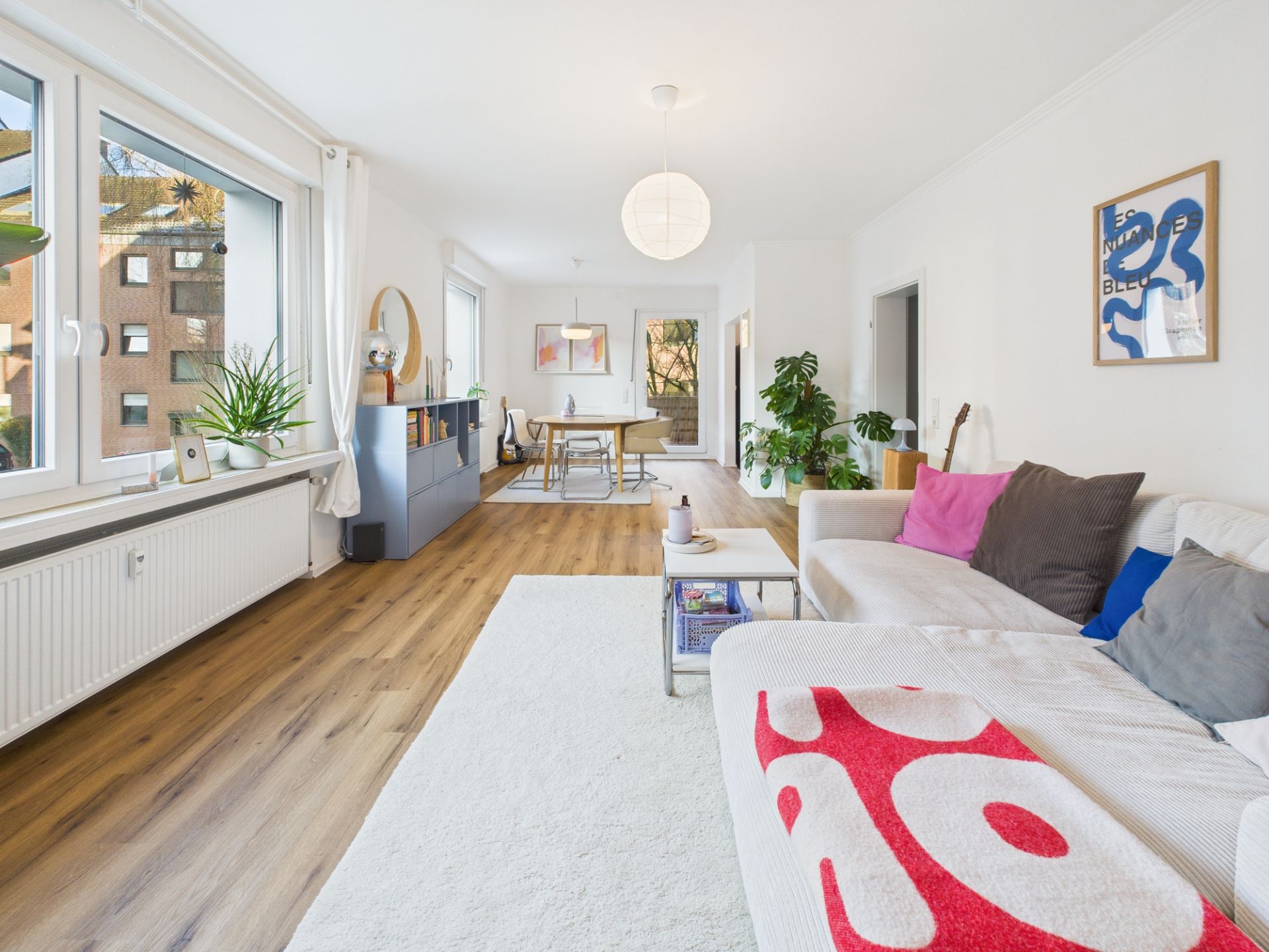 Helles Wohnzimmer mit Holzfußboden, Sofa, Pflanzen und großem Fenster, im Hintergrund Essbereich.