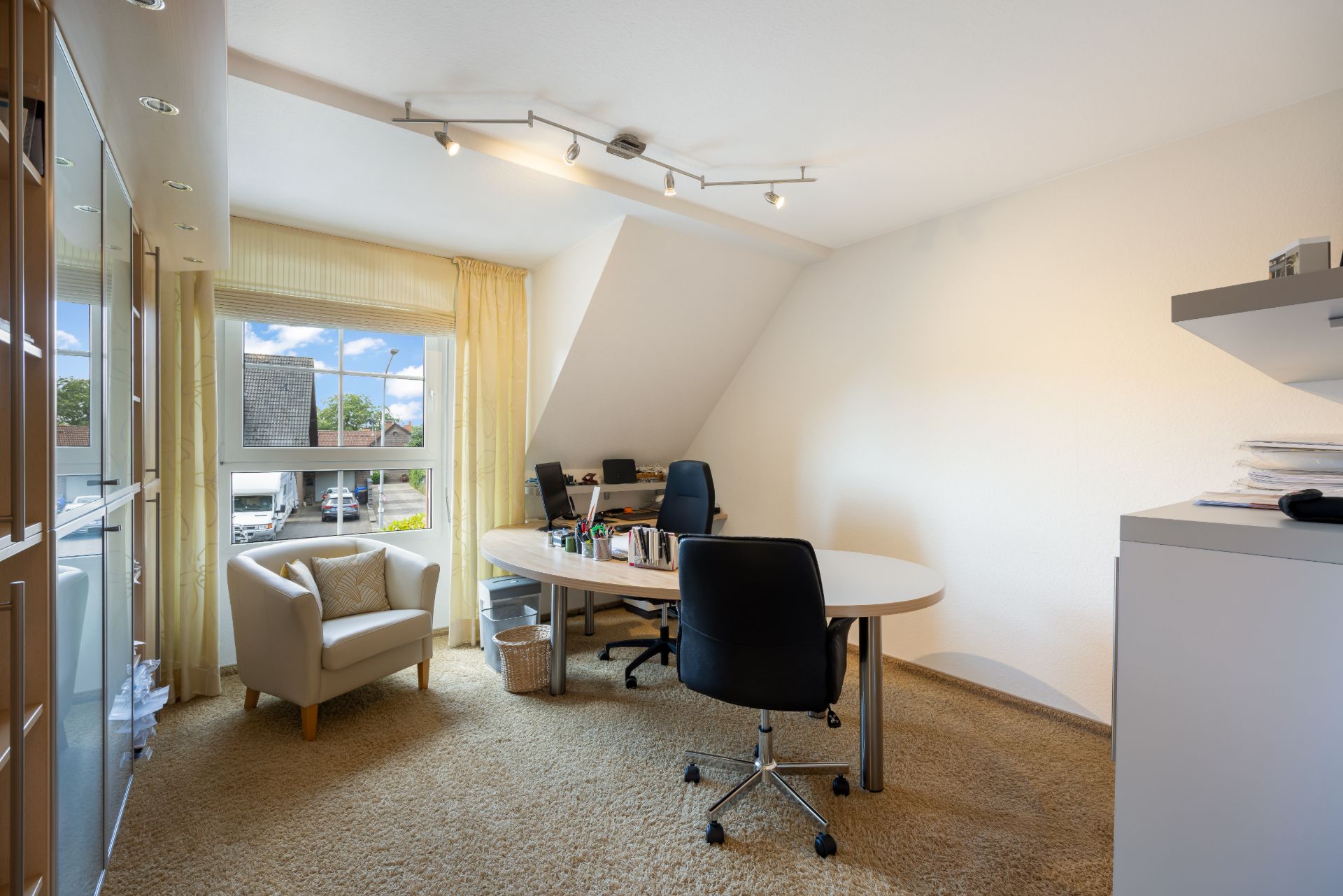Helles Arbeitszimmer mit Teppich, Schreibtisch, Stuhl und Sessel vor einem Fenster mit Gardinen.