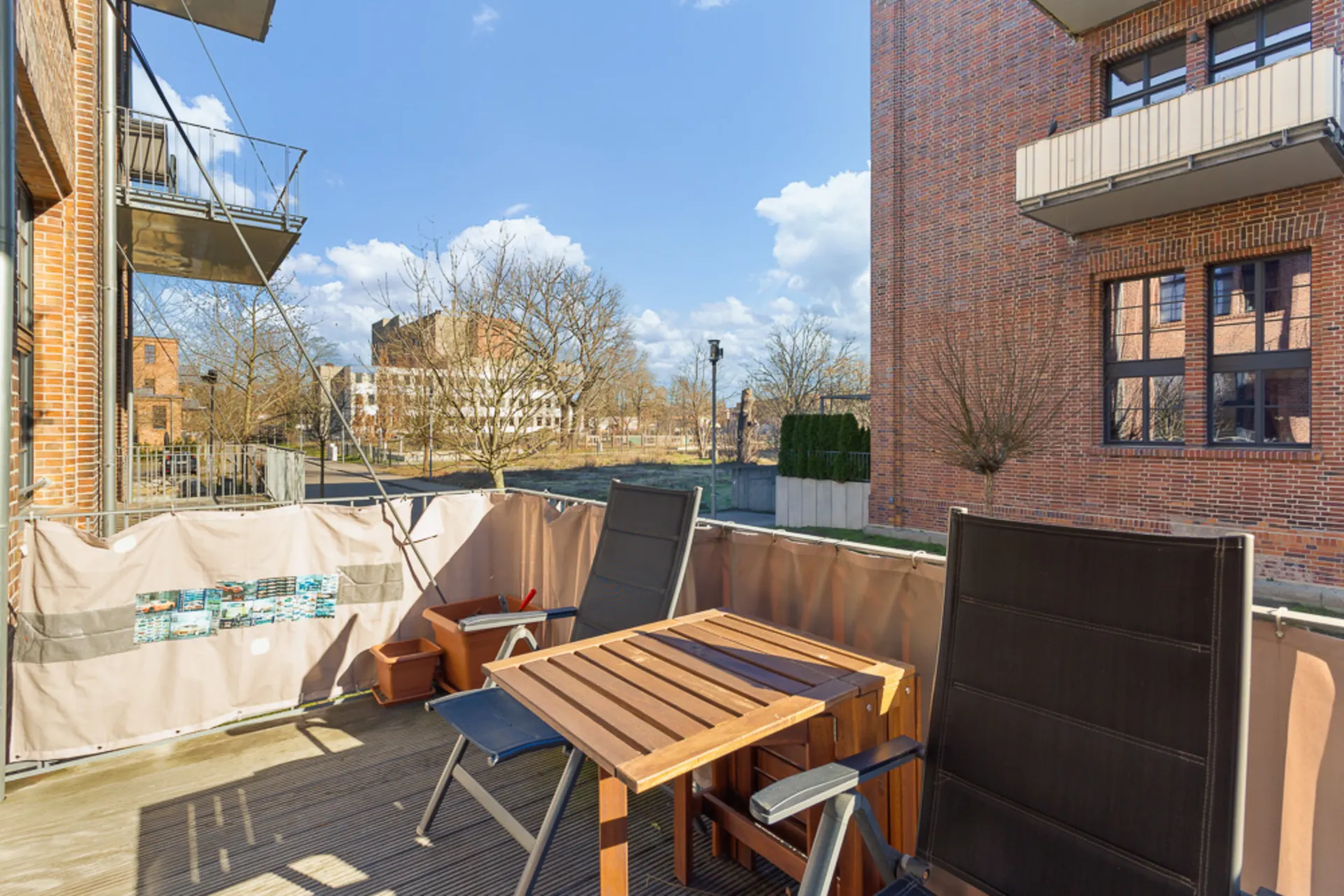 Balkon mit zwei Stühlen, Holztisch und Blumentöpfen, umgeben von Backsteinhäusern.