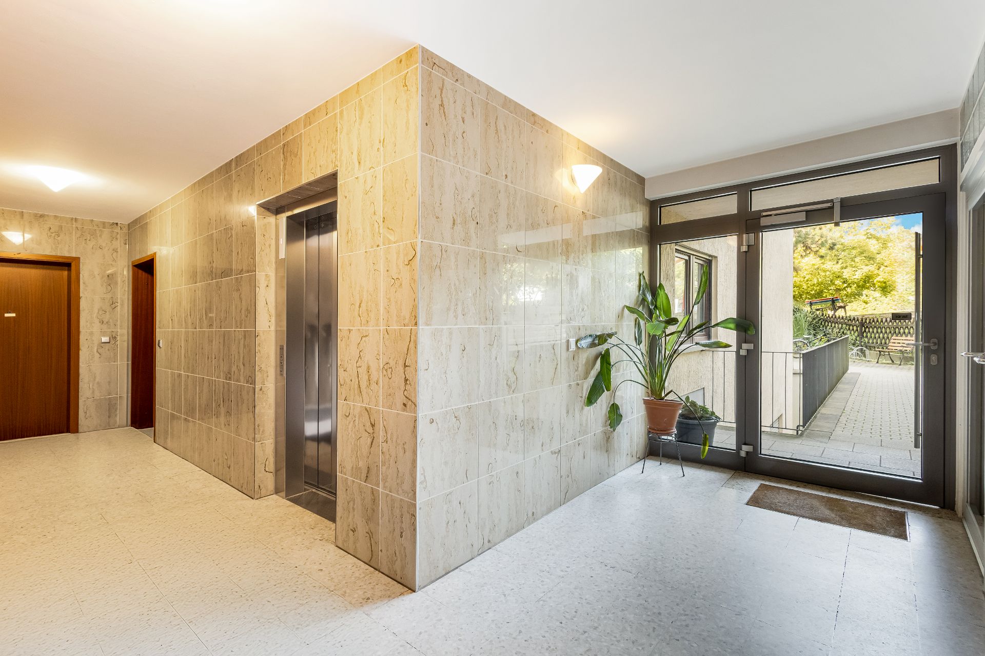 Heller Hausflur mit Fahrstuhl, Holztüren, Topfpflanze und Glastür zum Außenbereich.