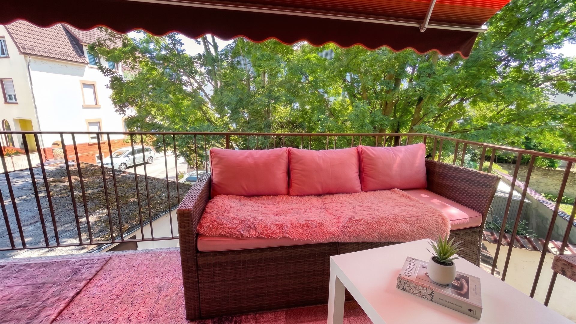 Balkon mit Sofa, rosa Kissen, kleinem Tisch und Blick auf Bäume und Nachbarhäuser.