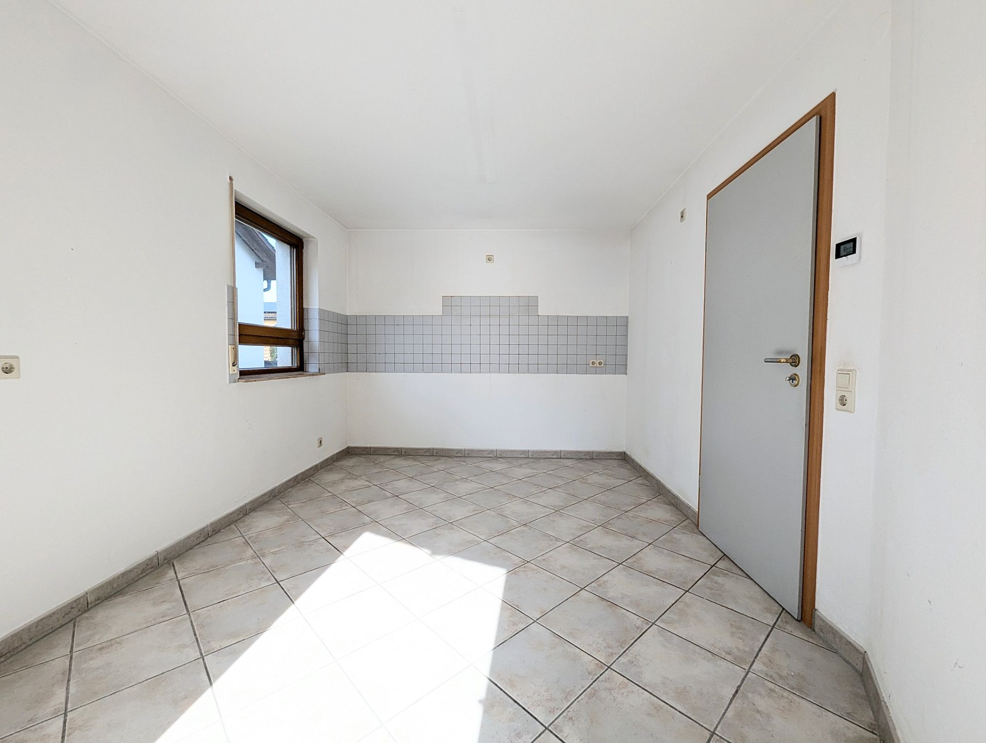 Leerer, heller Raum mit Fliesenboden, Fenster, Tür und gefliestem Wandbereich.
