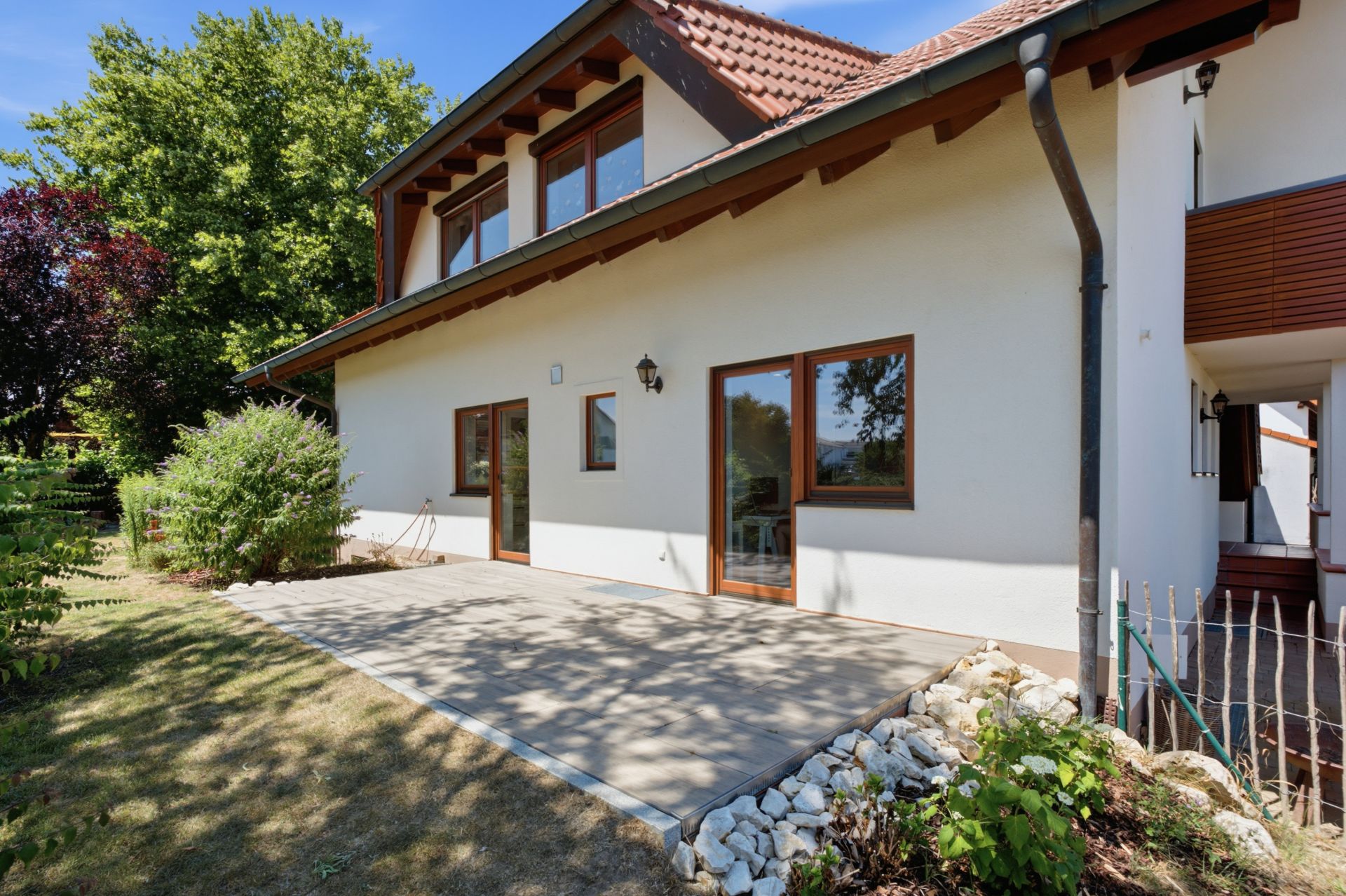Terrasse eines Wohnhauses mit Garten, großen Fenstern und Büschen vor der Hauswand.