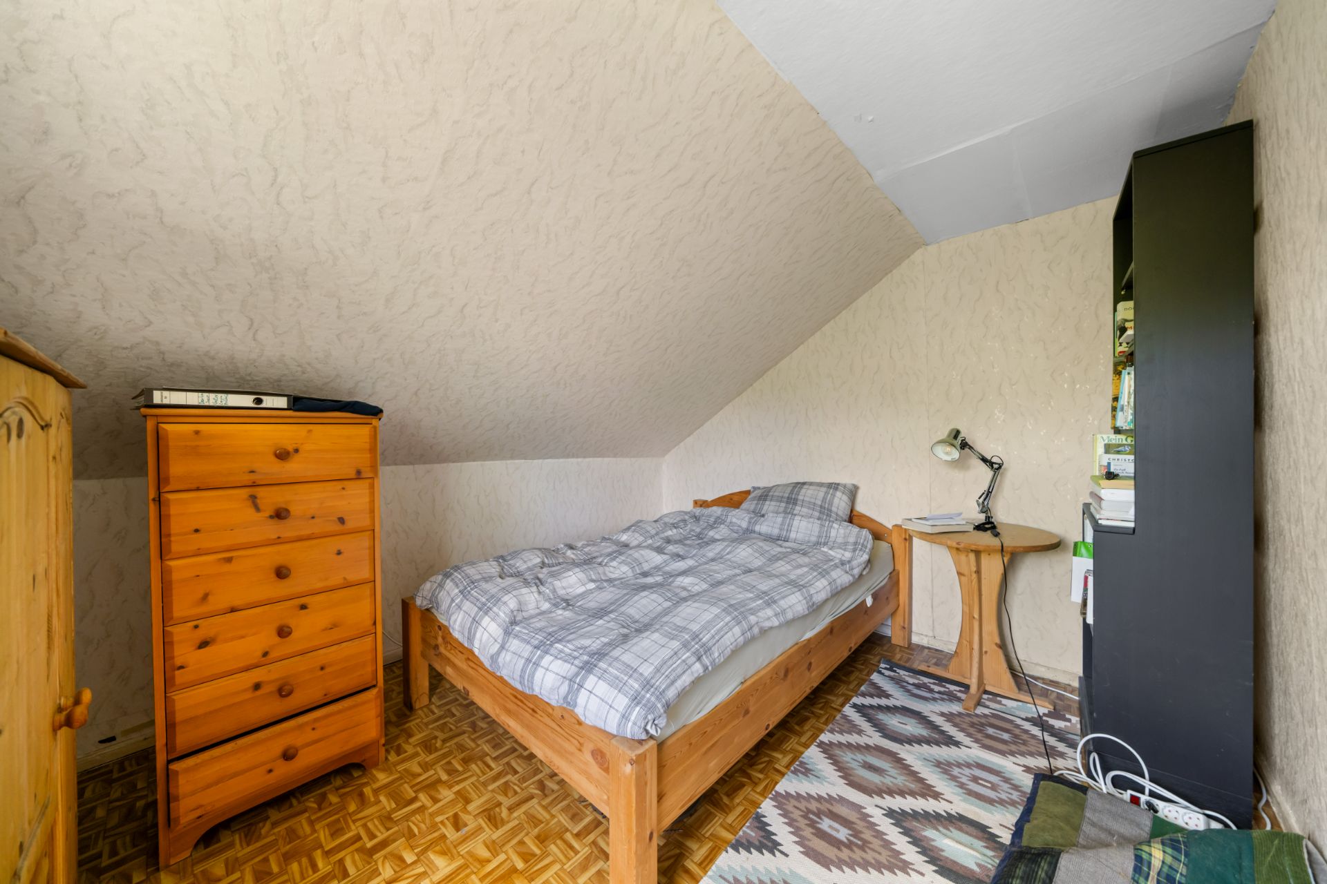 Kleines Schlafzimmer mit Einzelbett, Holzkommode, Nachttisch und Regal unter einer Dachschräge.