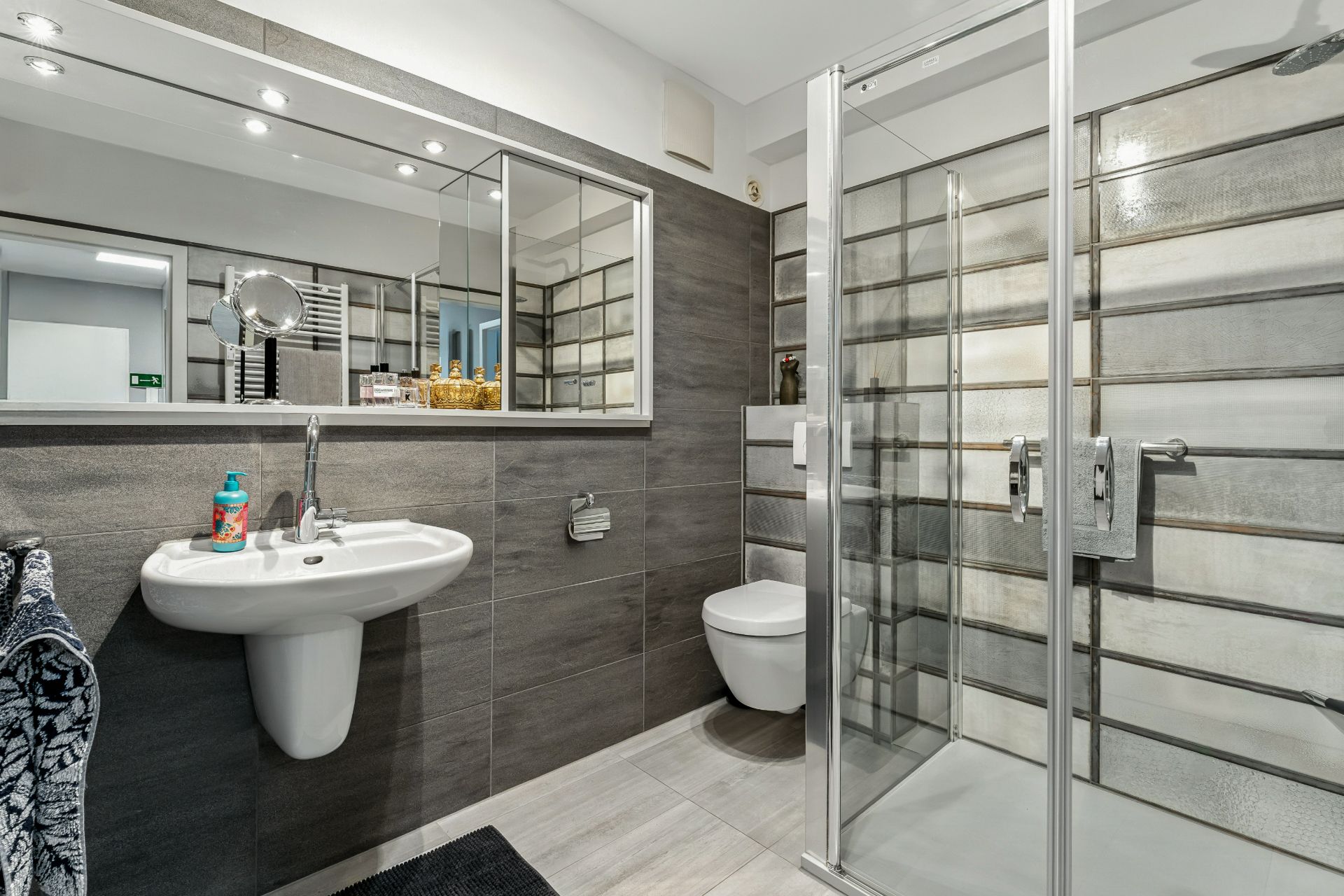 Modernes Badezimmer mit Dusche, Waschbecken, Spiegel, Wandtoilette und grauen Fliesen.