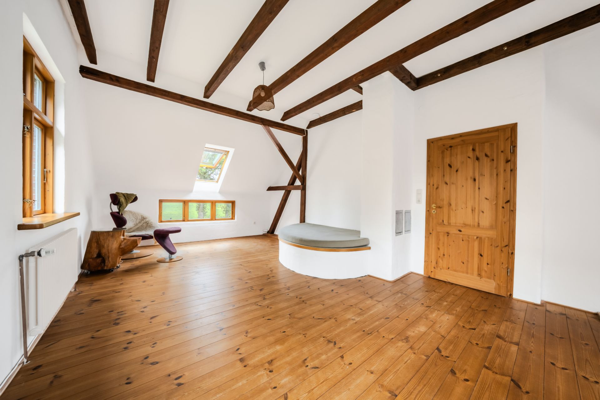 Helles Zimmer mit Holzboden, sichtbaren Deckenbalken, Holzfenstern und einer Holztür.