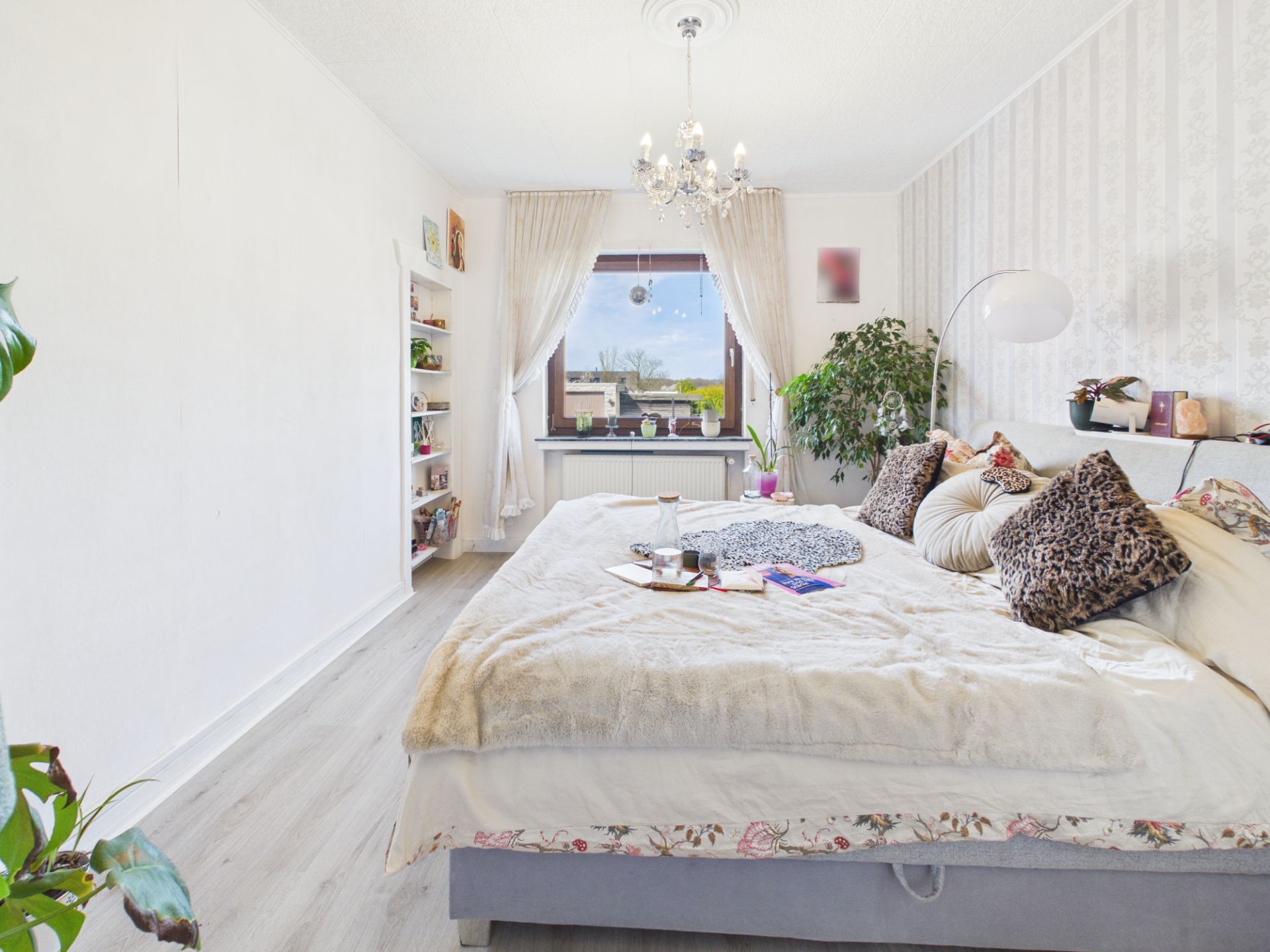 Helles Schlafzimmer mit großem Bett, vielen Kissen, Pflanzen und Fenster mit Blick nach draußen.
