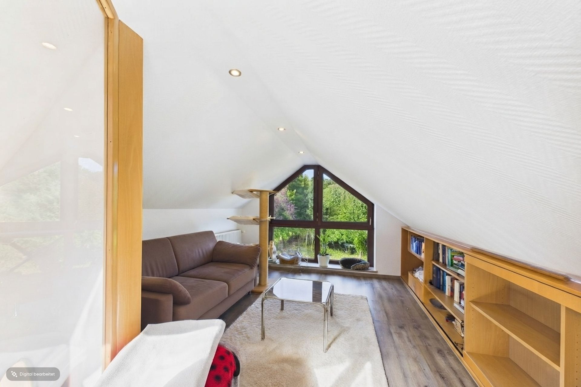 Heller Dachwohnraum mit Sofa, Couchtisch, Bücherregal und großen Fenstern mit Blick ins Grüne.