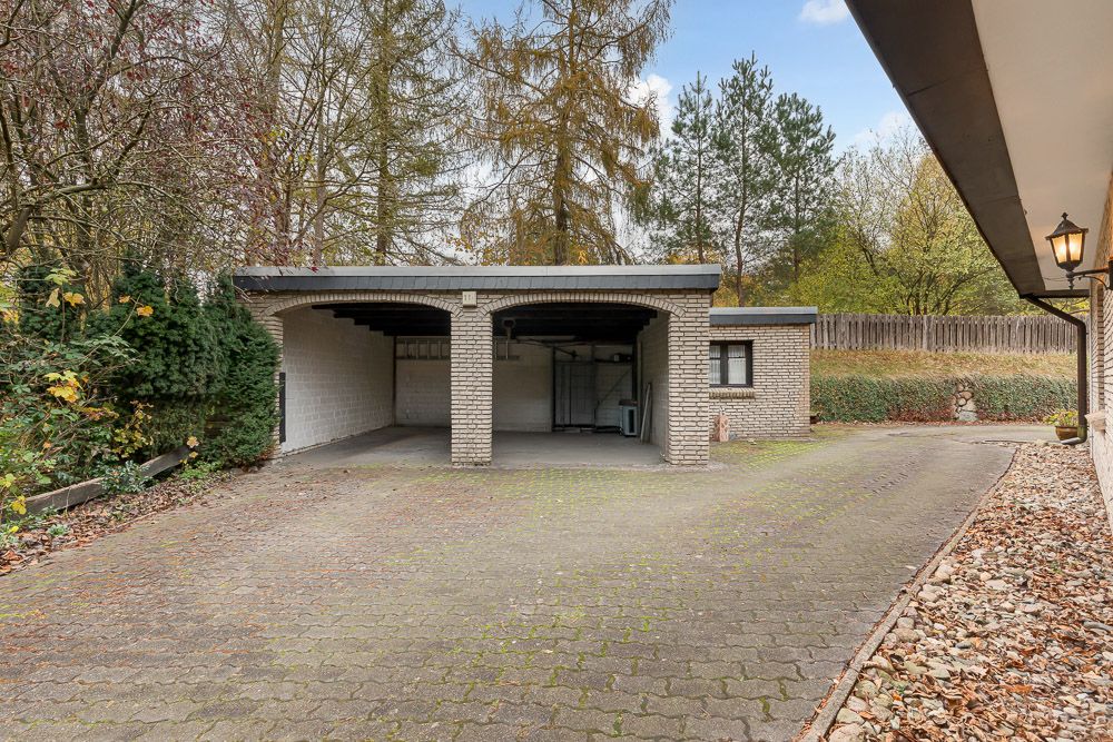 Überdachter Doppel-Carport aus Backstein mit gepflasterter Einfahrt, umgeben von Bäumen und Büschen.