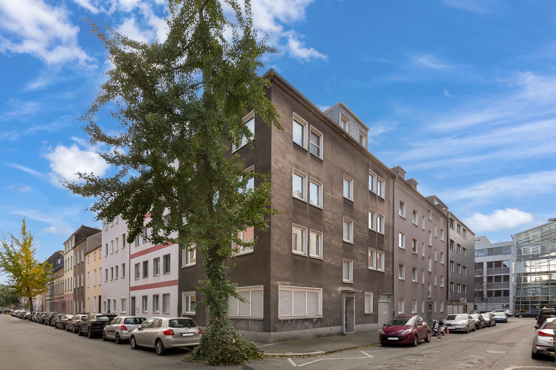 Mehrstöckiges Wohngebäude an einer Straßenecke mit geparkten Autos und Bäumen.