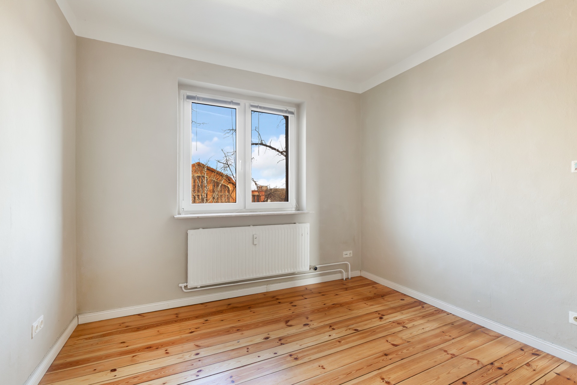 Leeres Zimmer mit Holzboden, hellem Anstrich und einem Fenster mit Blick nach draußen.