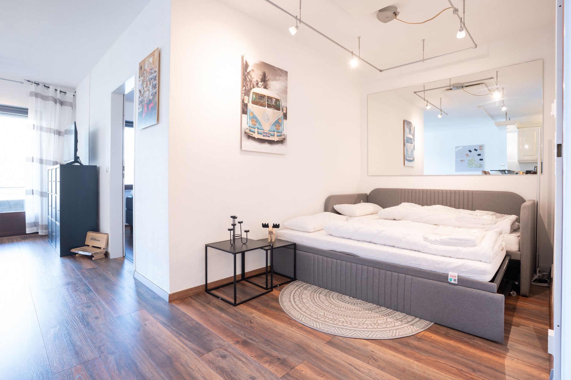 Helles Schlafzimmer mit grauem Bett, großem Spiegel, Holzboden und Wanddeko.