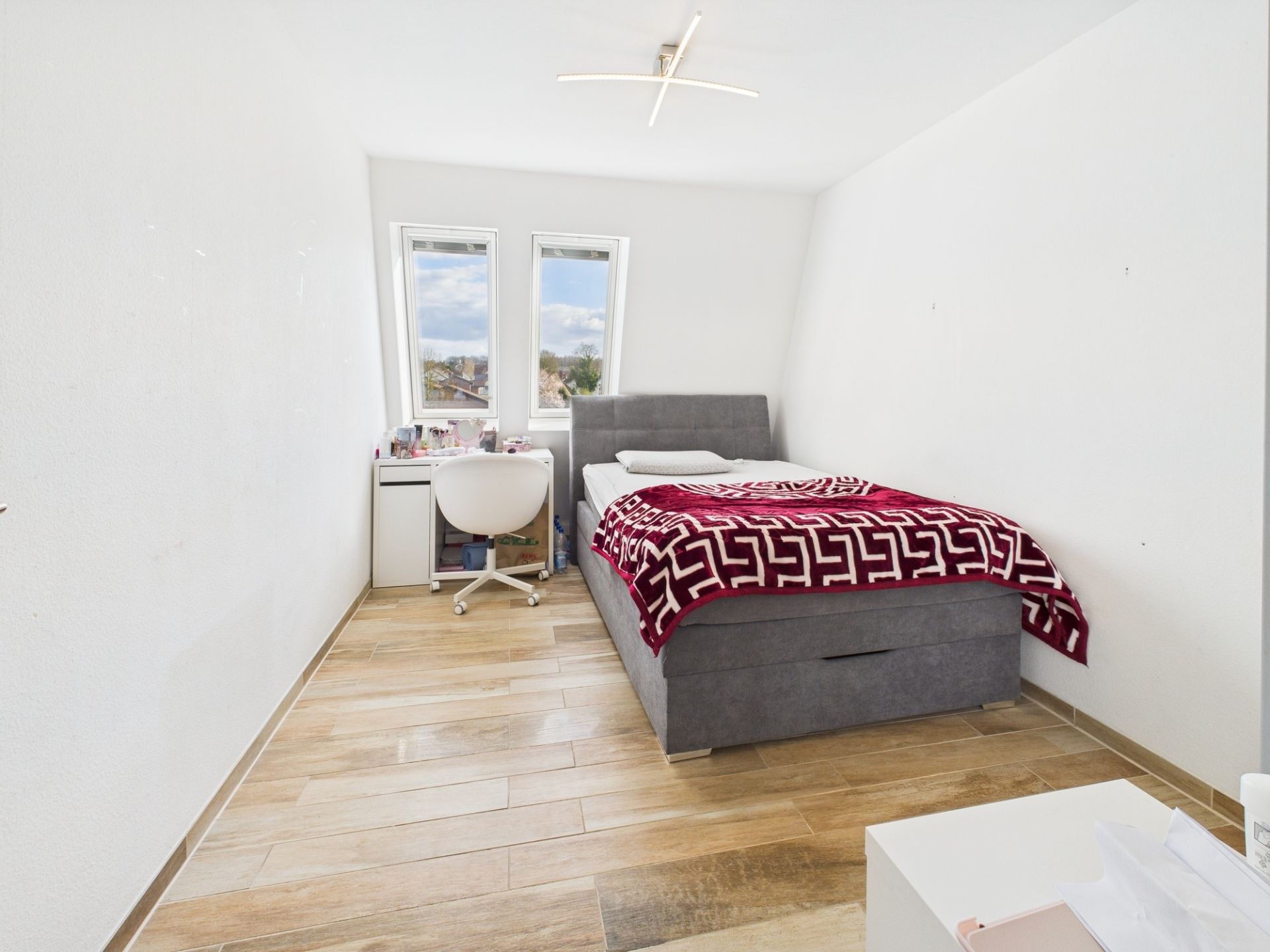 Helles Schlafzimmer mit grauem Bett, roter Decke, Schreibtisch und zwei Fenstern.