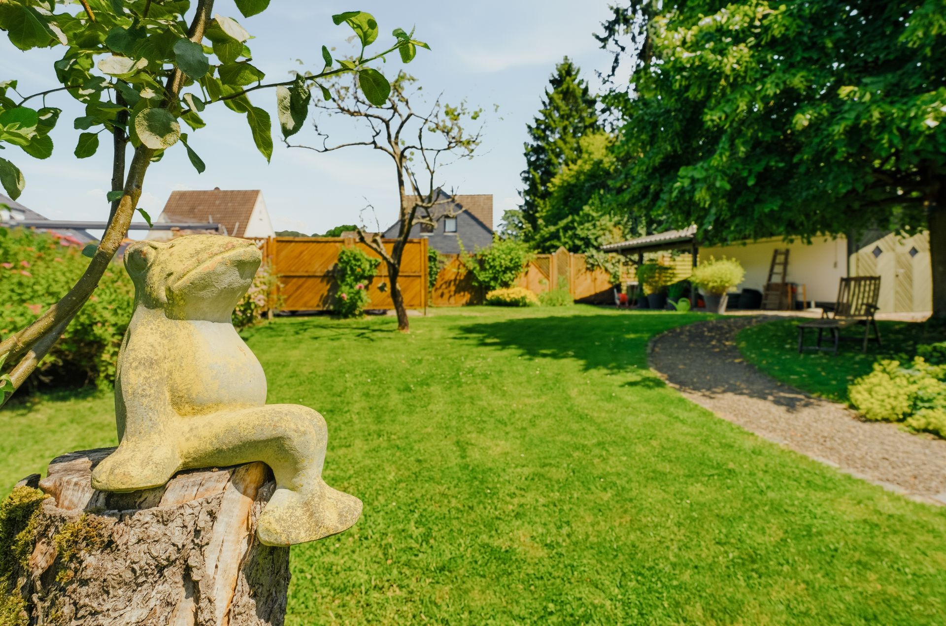 Garten mit Rasen, Baum, Gartenweg und Froschfigur auf einem Baumstumpf.