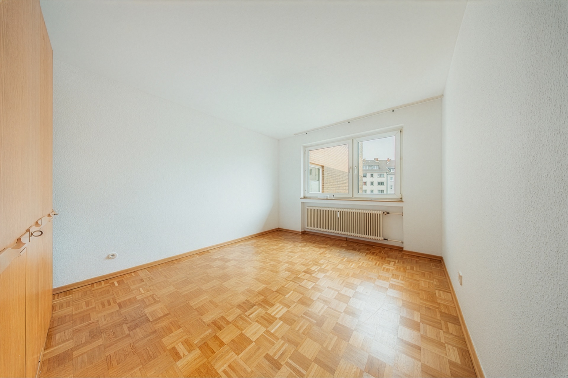 Leeres Zimmer mit Parkettboden, großem Fenster und Heizung.