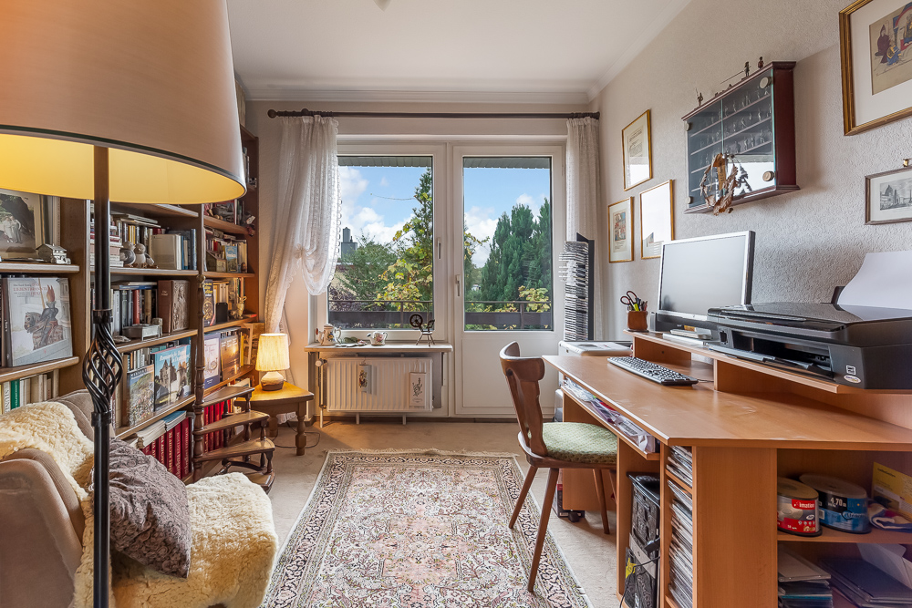Helles Arbeitszimmer mit Schreibtisch, Büchern, Sessel und großem Fenster mit Blick ins Grüne.