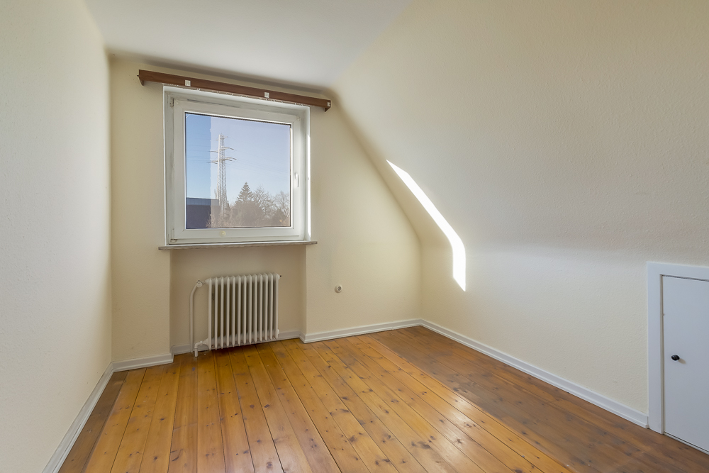Kleines leeres Zimmer mit Holzboden, Fenster, Heizkörper und Dachschräge.