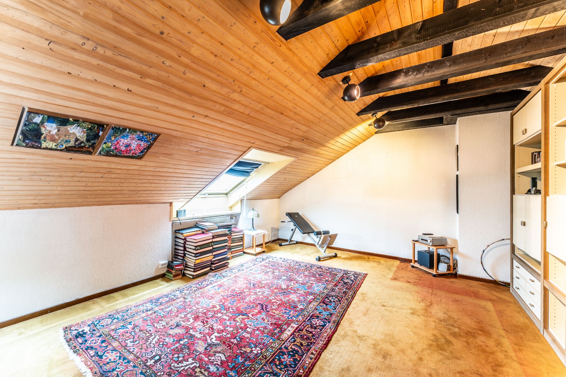Dachzimmer mit schräger Holzdecke, Teppich, Bücherstapeln, Regal und Hantelbank.
