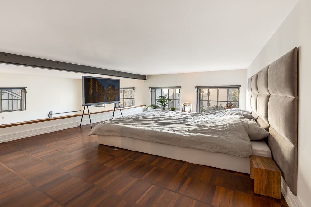Modernes Schlafzimmer mit großem Bett, Holzboden, Fernseher und mehreren Fenstern.
