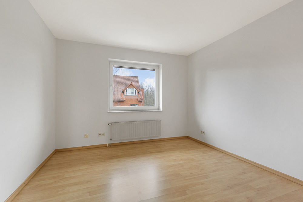 Leeres Zimmer mit hellem Holzfußboden, weißer Wand und Fenster mit Blick auf ein Haus.