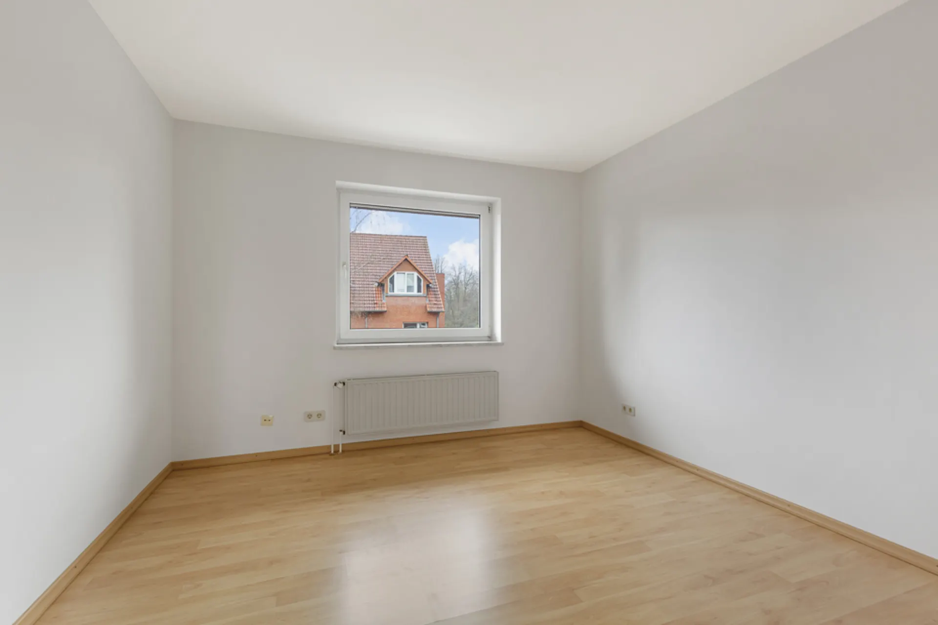 Leeres Zimmer mit hellem Holzfußboden, weißer Wand und Fenster mit Blick auf ein Haus.