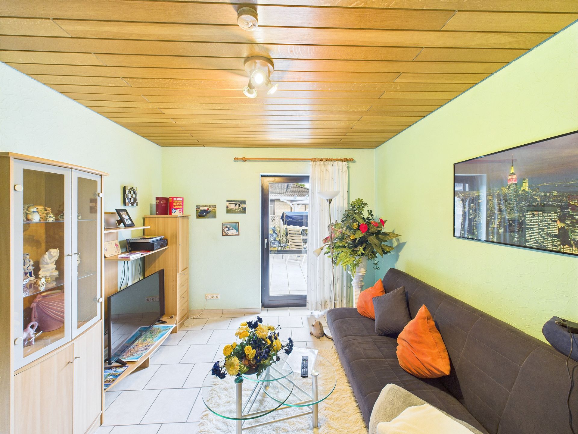 Wohnzimmer mit Sofa, Glastisch, TV, Blumen und Tür zum Balkon.