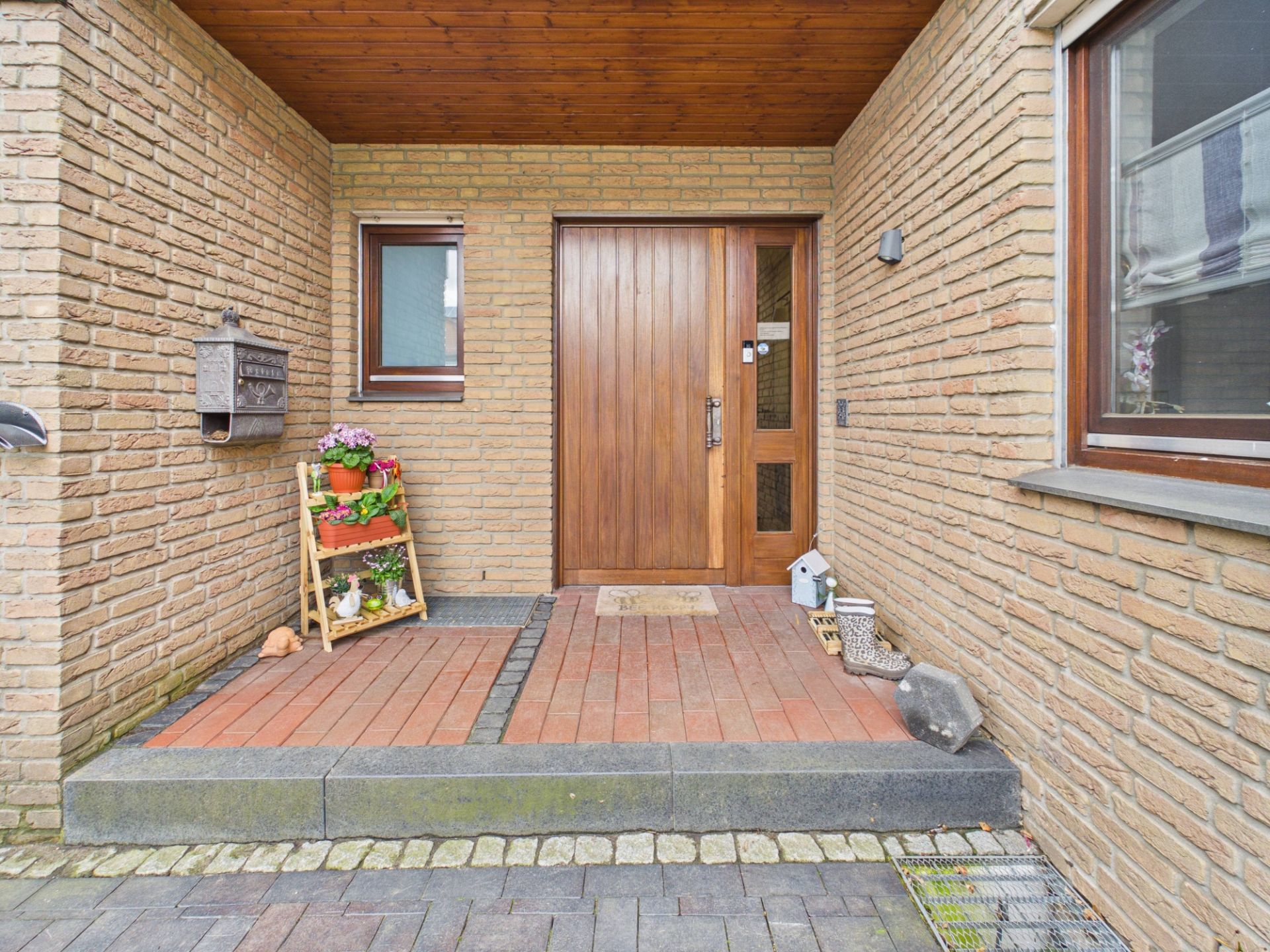 Eingangsbereich mit Holztür, kleiner Fenster, Blumentopf und Briefkasten an einer Backsteinwand.