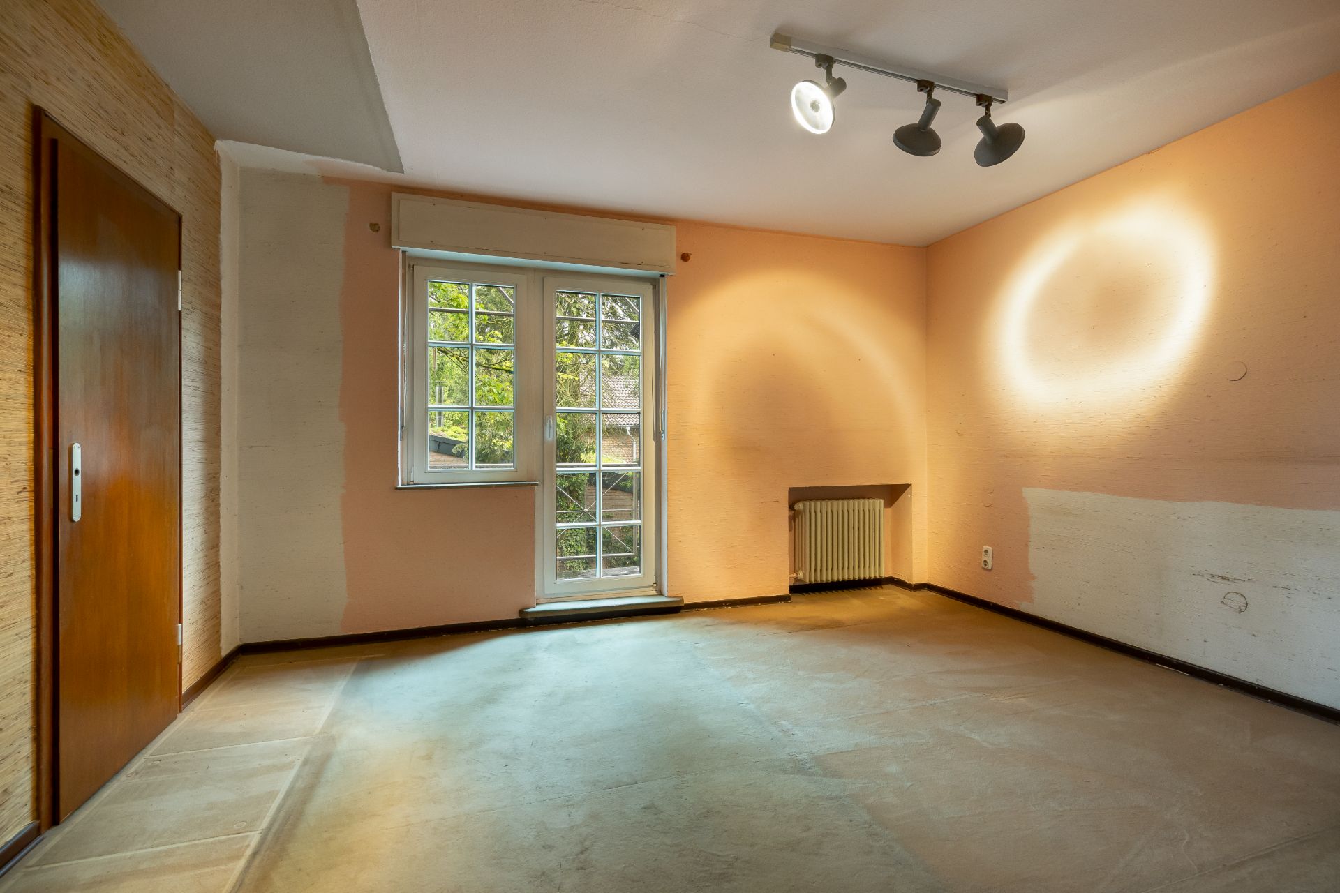Leerer Raum mit großem Fenster, Heizung und heller Beleuchtung an der Decke.