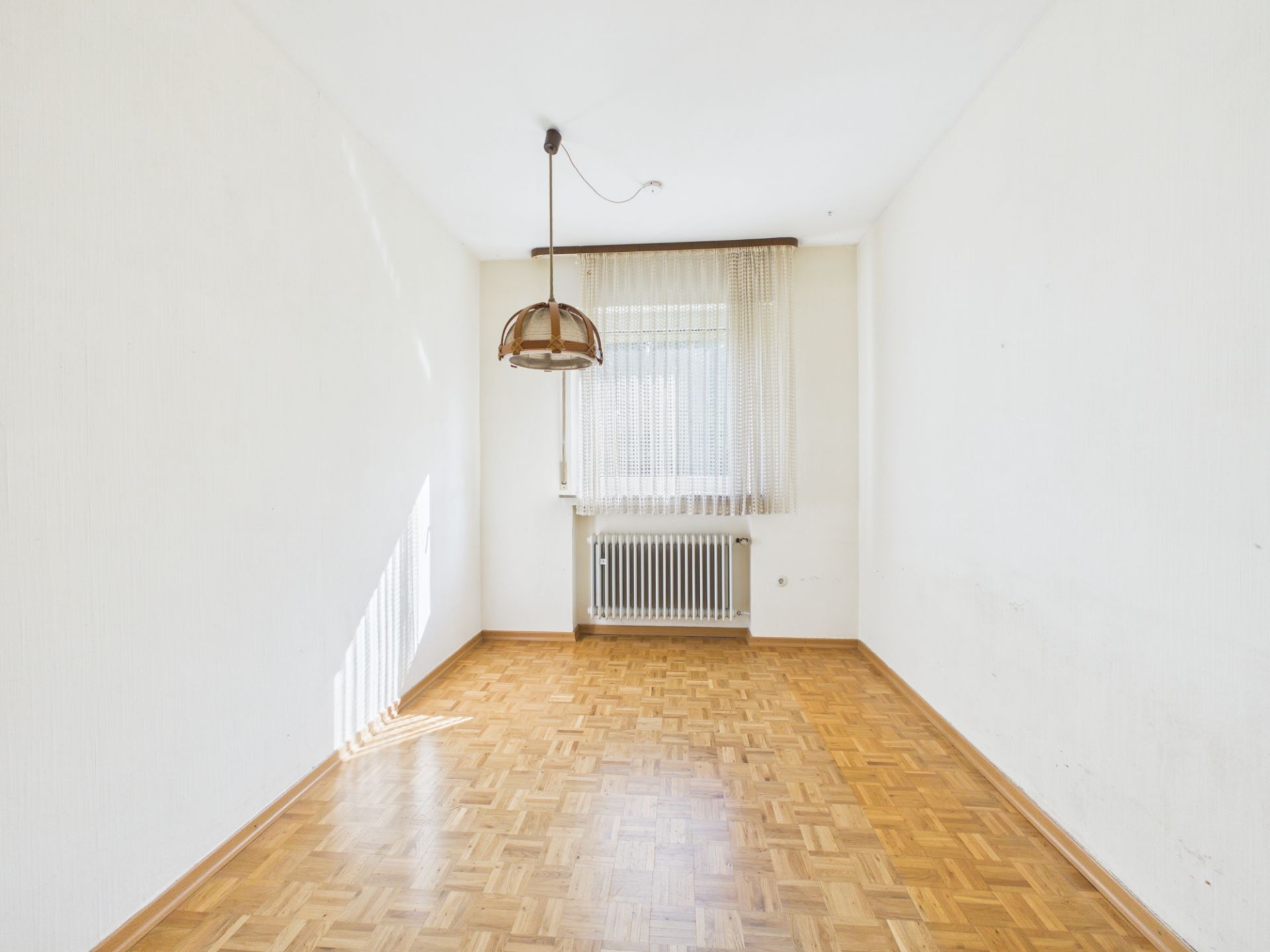 Leeres Zimmer mit Parkettboden, Hängelampe, Fenster mit Gardine und Heizkörper.