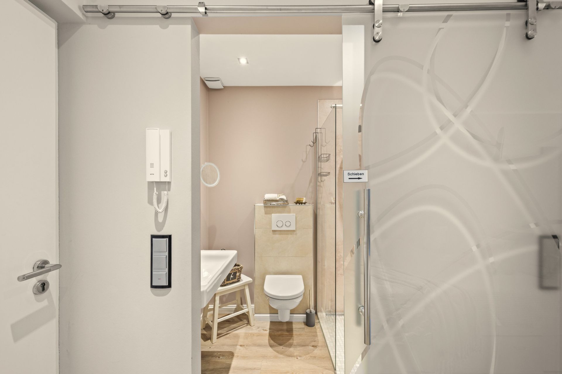 Helles Badezimmer mit WC, Dusche, Waschbecken und Hocker, Blick durch eine Schiebetür.