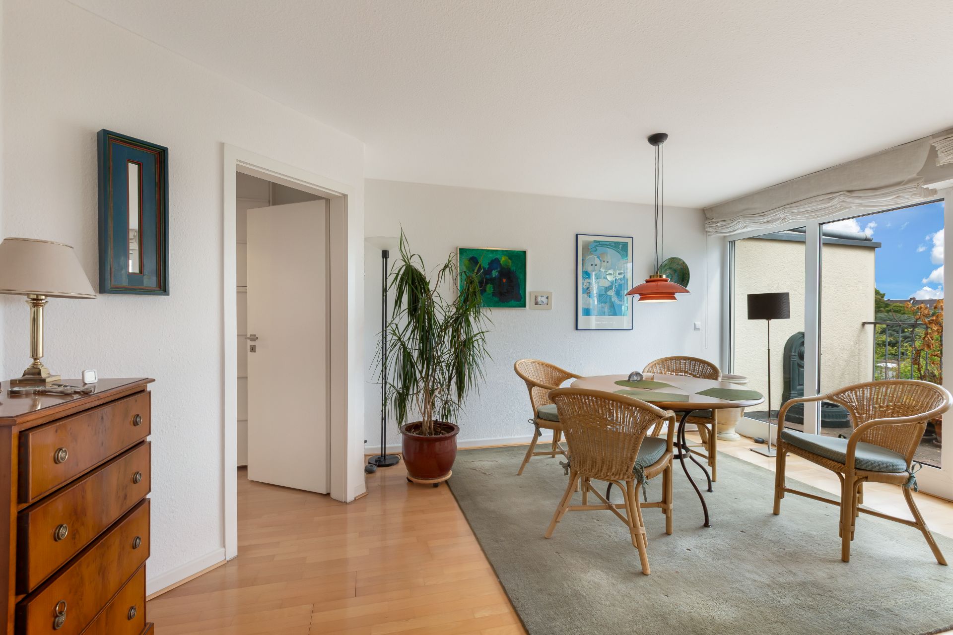 Essbereich mit Holztisch, vier Stühlen, großer Pflanze, Kommode und Fensterfront mit Blick nach draußen.