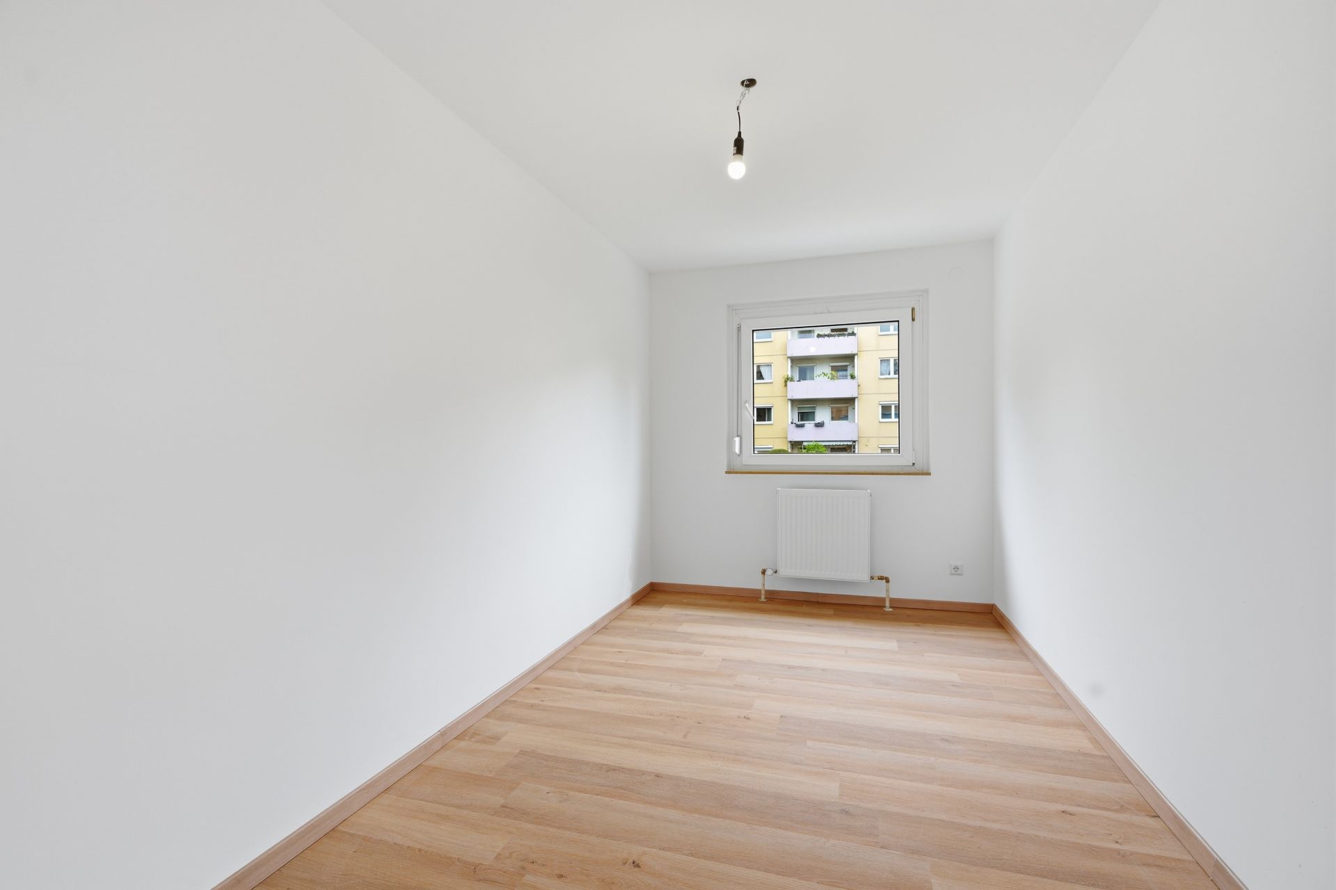 Leerer, schmaler Raum mit hellem Holzboden, weißer Wand und einem Fenster mit Blick nach draußen.
